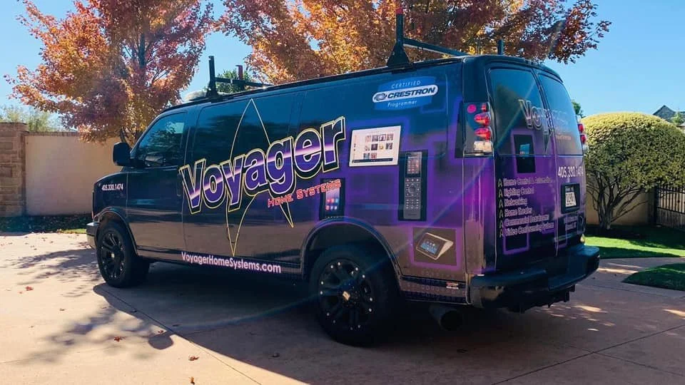 A dark blue Voyager Home Systems service van parked on a driveway, with fall trees in the background.