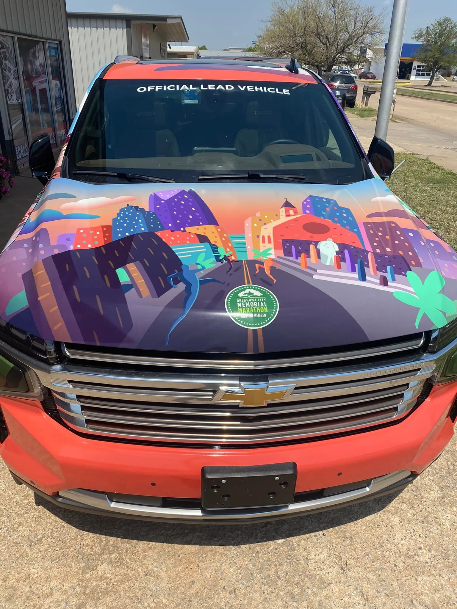 Front view of a Chevrolet vehicle with a colorful cityscape illustration on the hood, featuring skyscrapers, a bridge, and running figures, along with a green sticker for the Oklahoma City Memorial Marathon.
