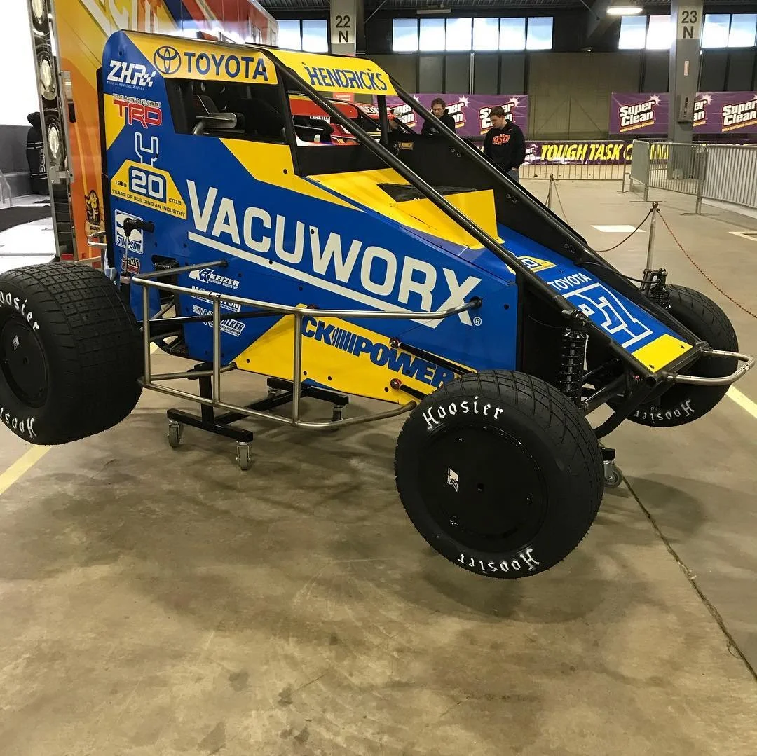 A blue and yellow sprint car with large Hoosier tires, displayed indoors at an event or exhibition, with sponsorship stickers and branding including Vacuum Wory, Toyota, and others.