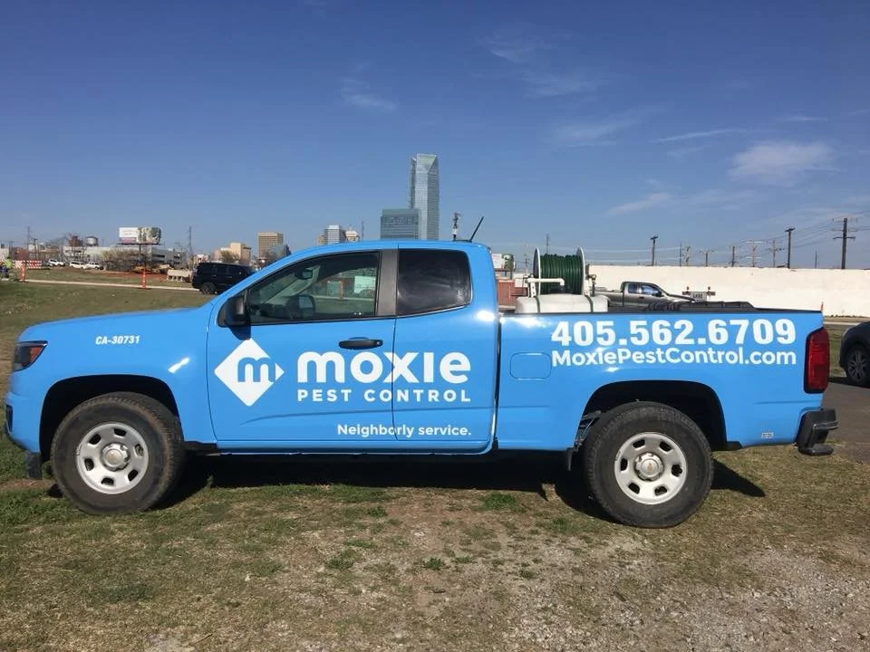 Blue Moxie Pest Control truck parked on grass with city Skyline in the background.