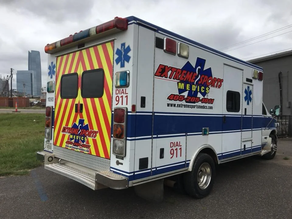 Ambulance emergency vehicle with 'Extreme Sports Medics' logos and contact information parked outdoors with city buildings and cloudy sky in the background.