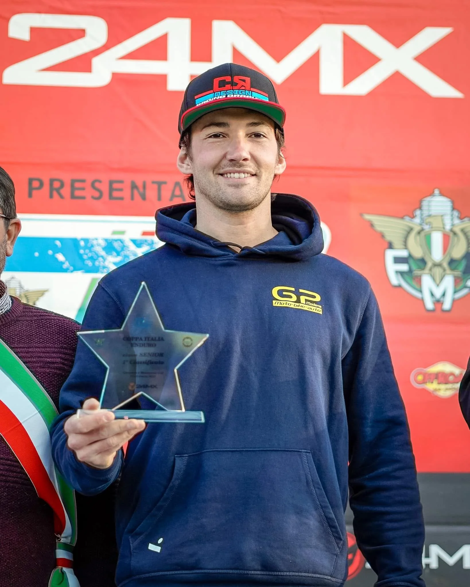 Presidente GP Modena Pietro Pini con cappellino e felpa blu tiene in mano un trofeo a forma di stella, sorridendo, davanti a uno sfondo rosso con scritte e loghi di motori motociclistici.