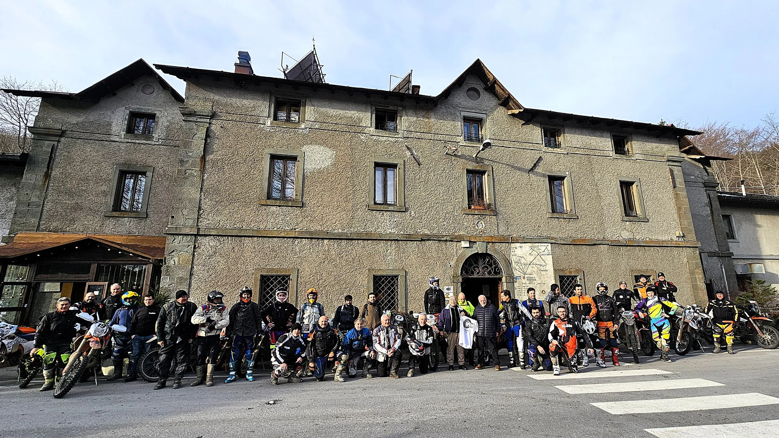 Gruppo di motociclisti con moto davanti a un edificio antico in pietra con finestre e un arco d'ingresso.