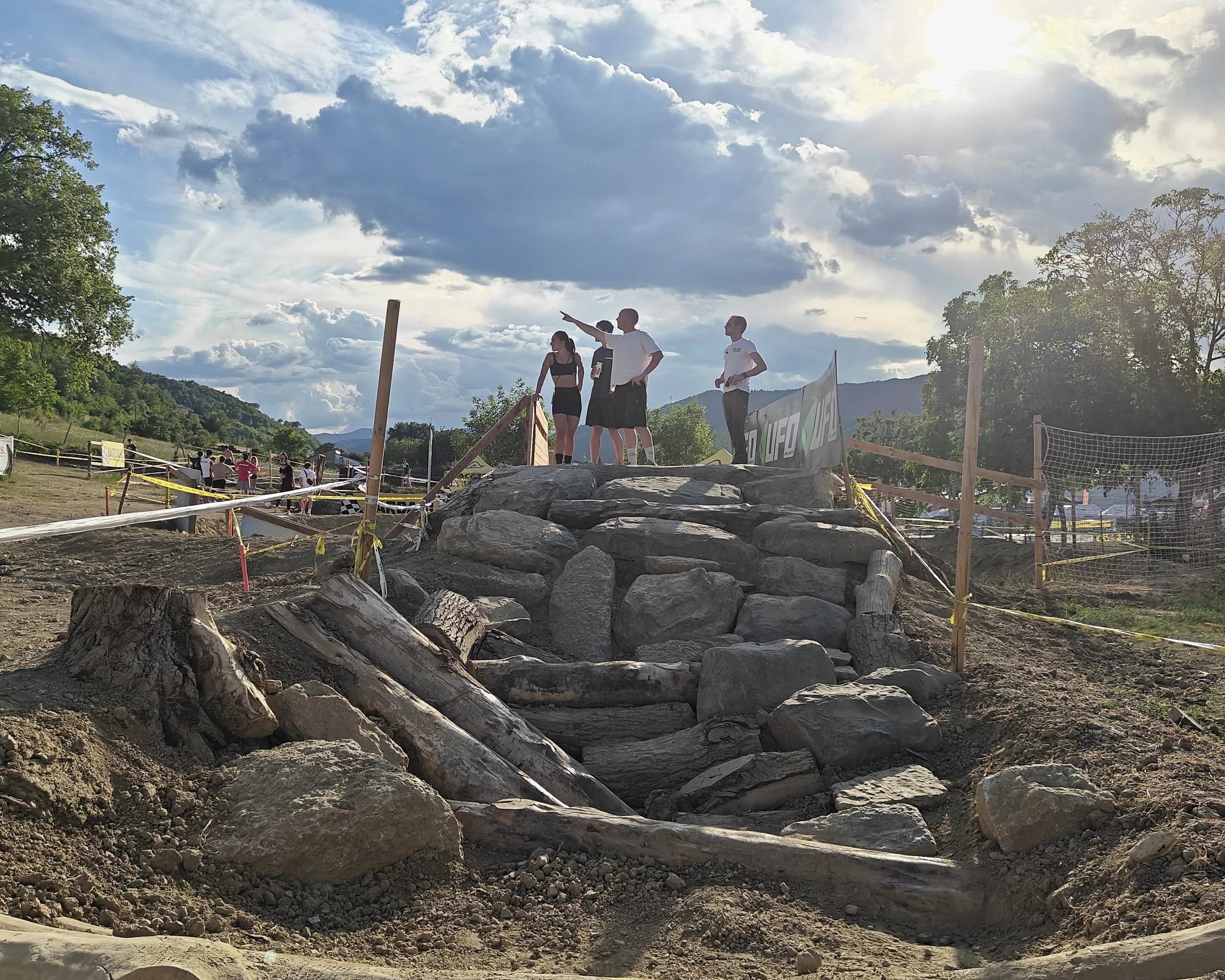 Gruppo di persone che discute su una struttura fatta di pietre e tronchi, con un cielo nuvoloso sullo sfondo, apparentemente durante un evento all'aperto.