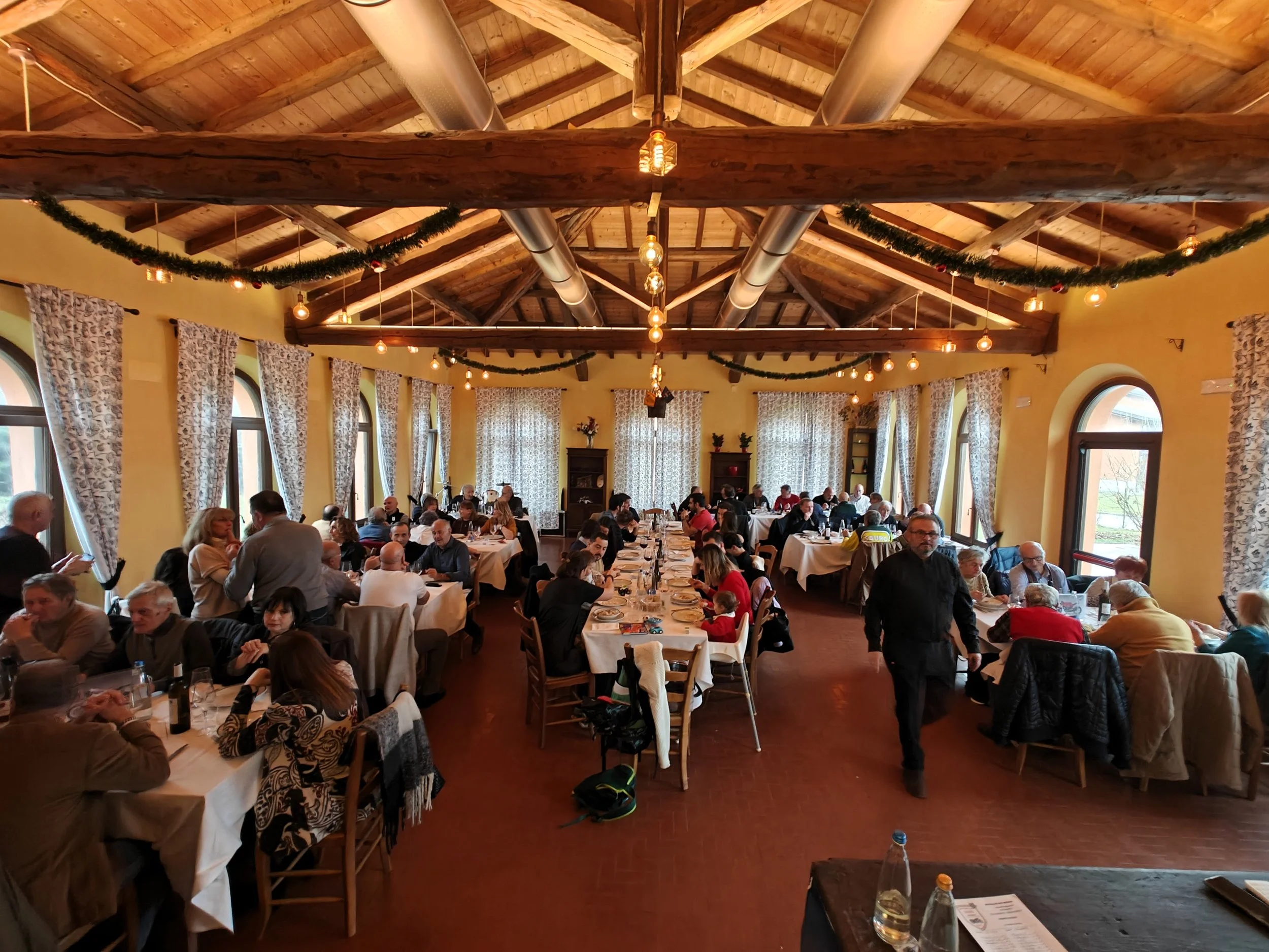 Interno di un ristorante affollato durante una festa, con persone sedute a tavoli rettangolari decorati con tovaglie beige, illuminazione calda e decorazioni natalizie come ghirlande verdi. La sala ha pareti gialle, grandi finestre con tende floreali e un soffitto in legno con travi a vista.