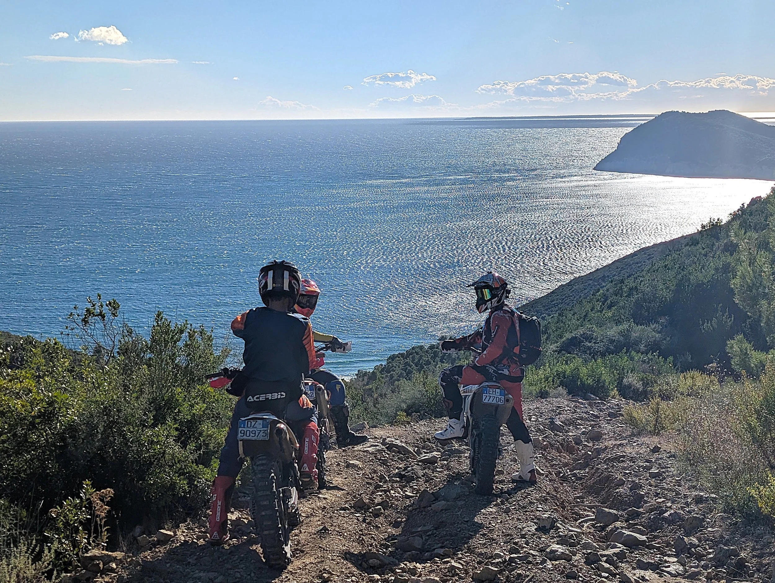 Tre motociclisti in tuta da motocross con caschi su un sentiero di montagna vicino al mare, con una vista sull'oceano e una piccola isola all'orizzonte.
