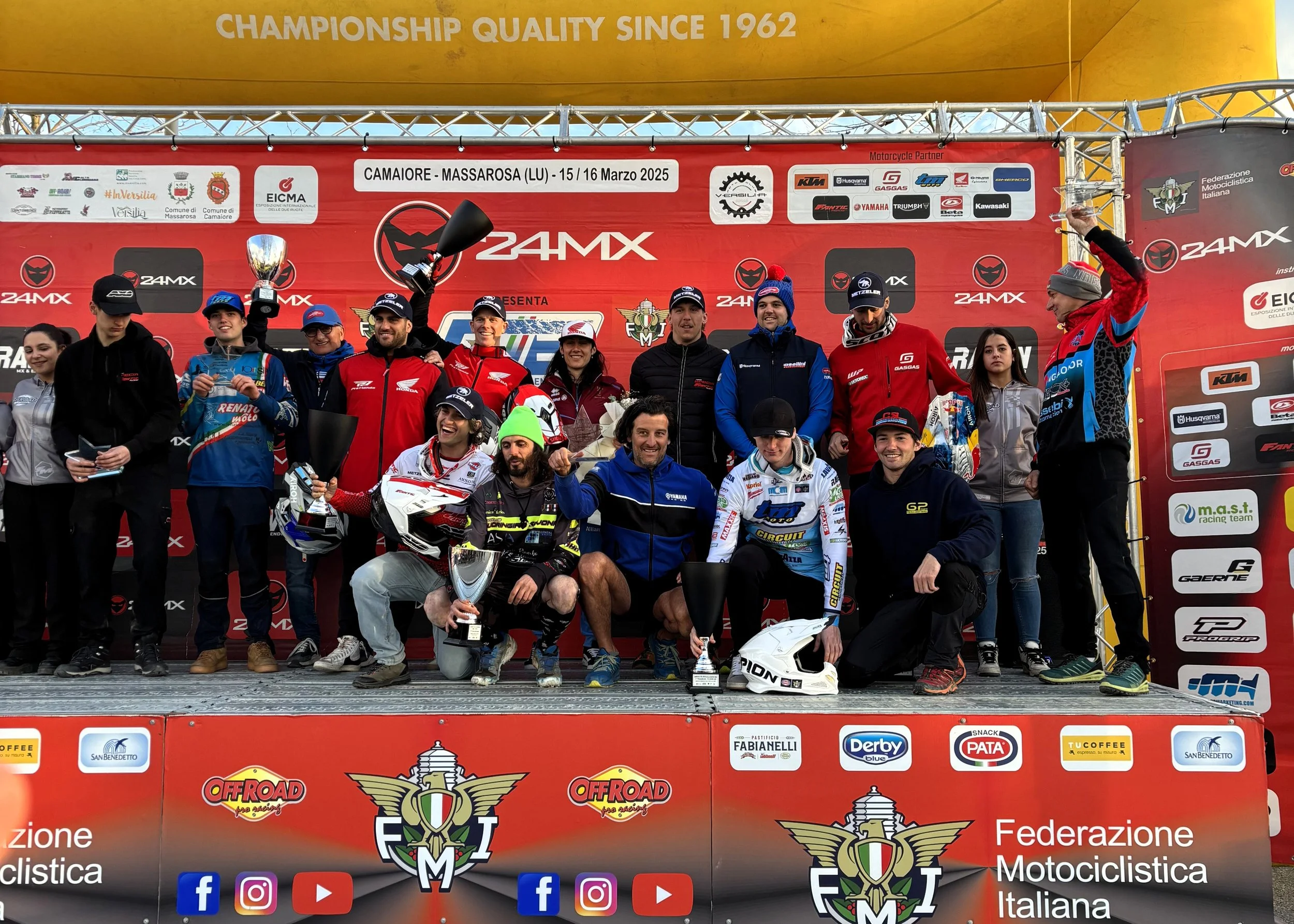 Gruppo di piloti e team di motociclismo in piedi sul podio, alcuni con trofei, con sfondo rosso e loghi di eventi e sponsor.