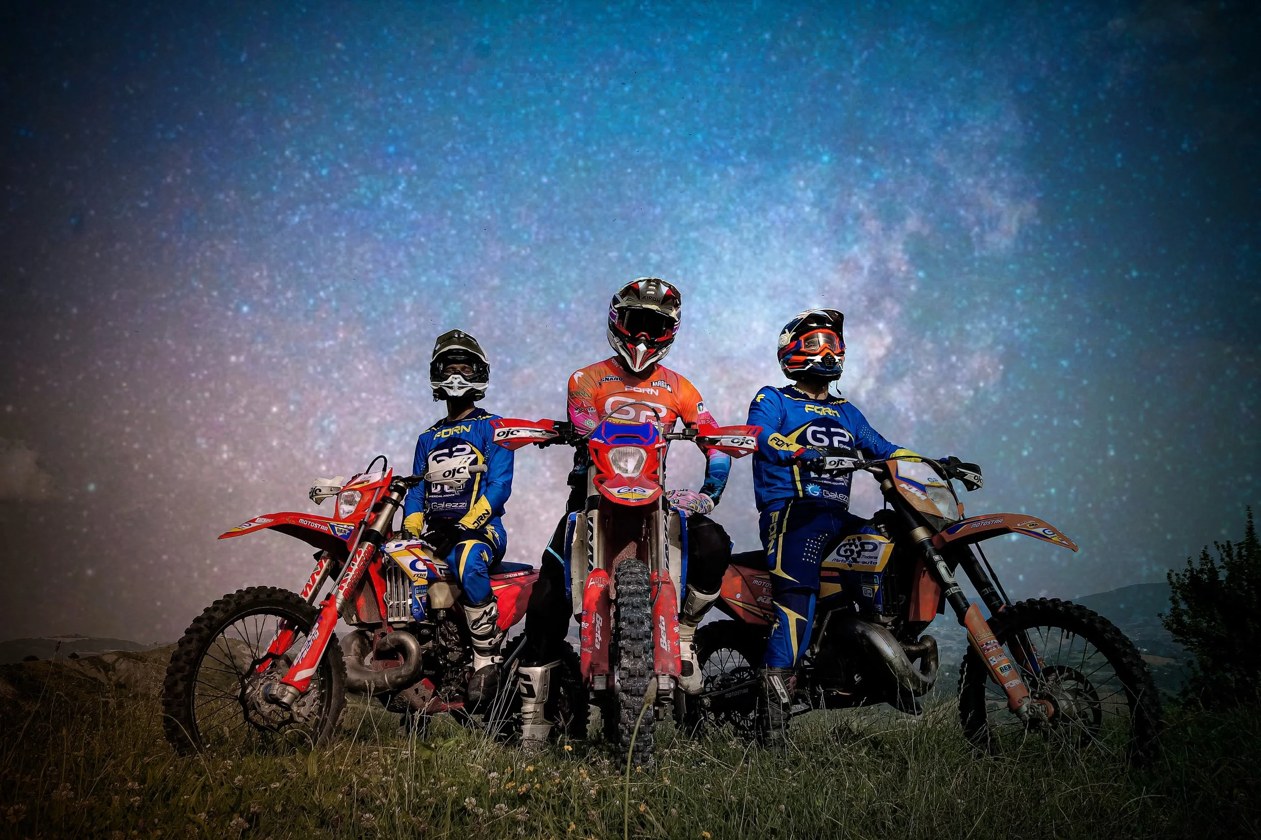 Tre enduristi in tenuta colorata con caschi e visiere, in piedi con le moto su un prato, sotto un cielo stellato notturno.