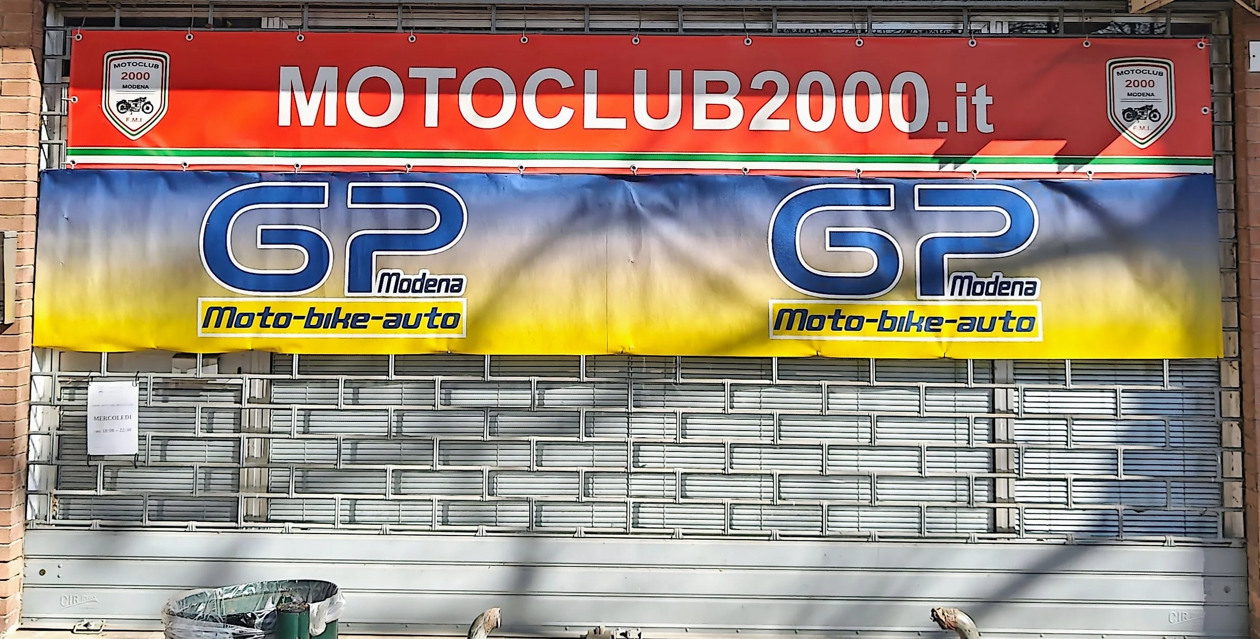 Vetrina con cartelloni pubblicitari per Moto Club 2000 Modena e GP Modena. L'insegna sopra è rossa con testo bianco. La pubblicità sotto è colorata con sfumature di blu, bianco e giallo. Si tratta della sede del motoclub GP modena
