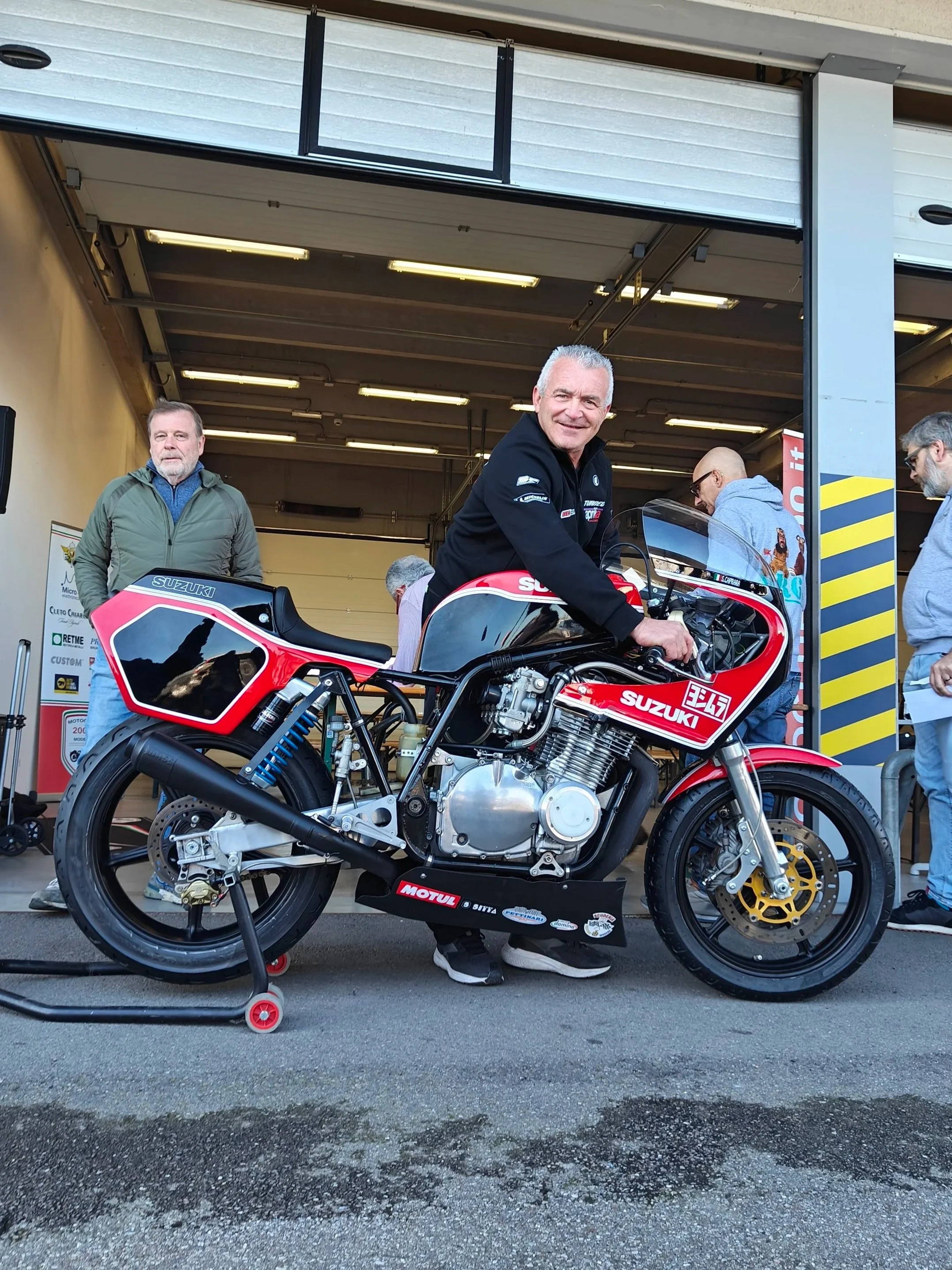 Sandro Caprara ex Presidente Motoclub 2000 su una motocicletta da corsa Suzuki nel box di un circuito, con altri uomini che si preparano intorno. Il motociclista sorride e guarda verso la fotocamera.
