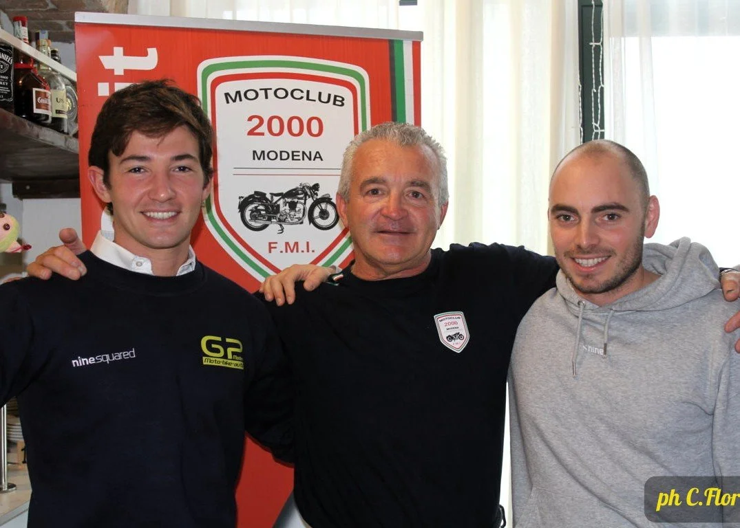 Tre uomini sorridenti si abbracciano davanti a un banner del Motoclub 2000 di Modena, con un'illustrazione di una moto, in un ambiente interno con scaffali di bottiglie e decorazioni.