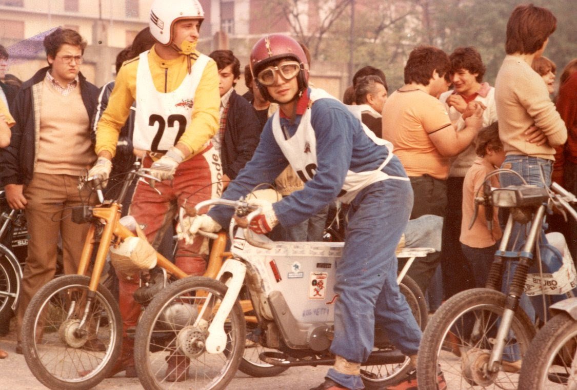 Giovani partecipanti a una gara di ciaocross, alcuni sullo sfondo e altri in primo piano con maglie numerate, indossano caschi e abbigliamento da corsa, circondati da un pubblico di spettatori.