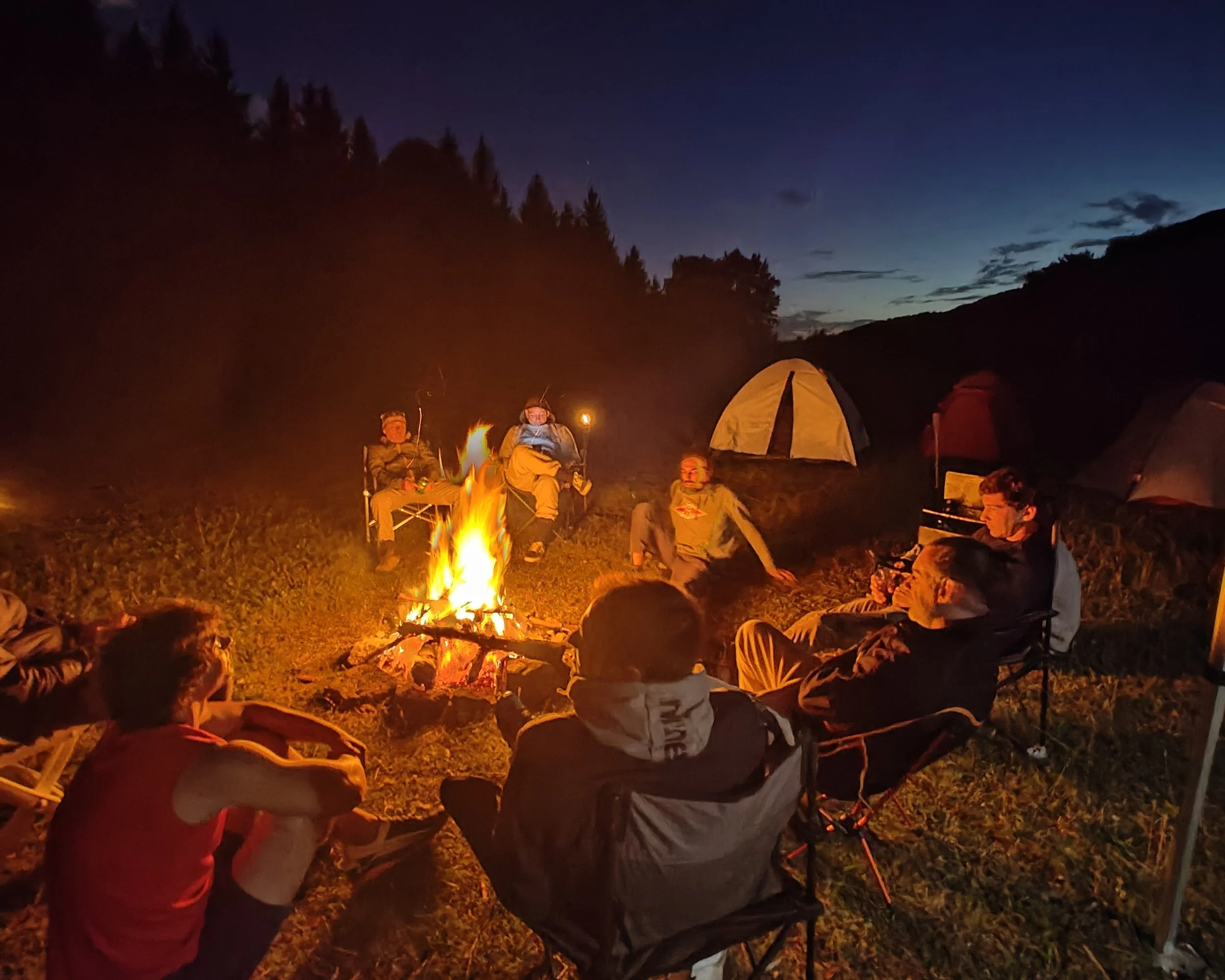 Gruppo di persone sedute intorno a un falò in campeggio durante la sera, con tende illuminate sullo sfondo e un cielo notturno.