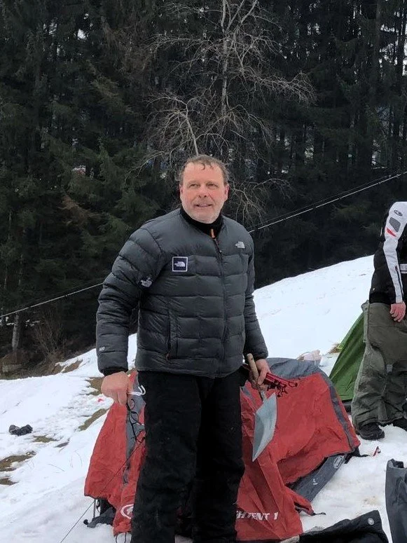 Carlo Rinaldi responsabile area moto storiche con giacca nera in stile invernale, in un'area nevosa con tende e alberi sullo sfondo, durante un'escursione o campeggio in montagna.