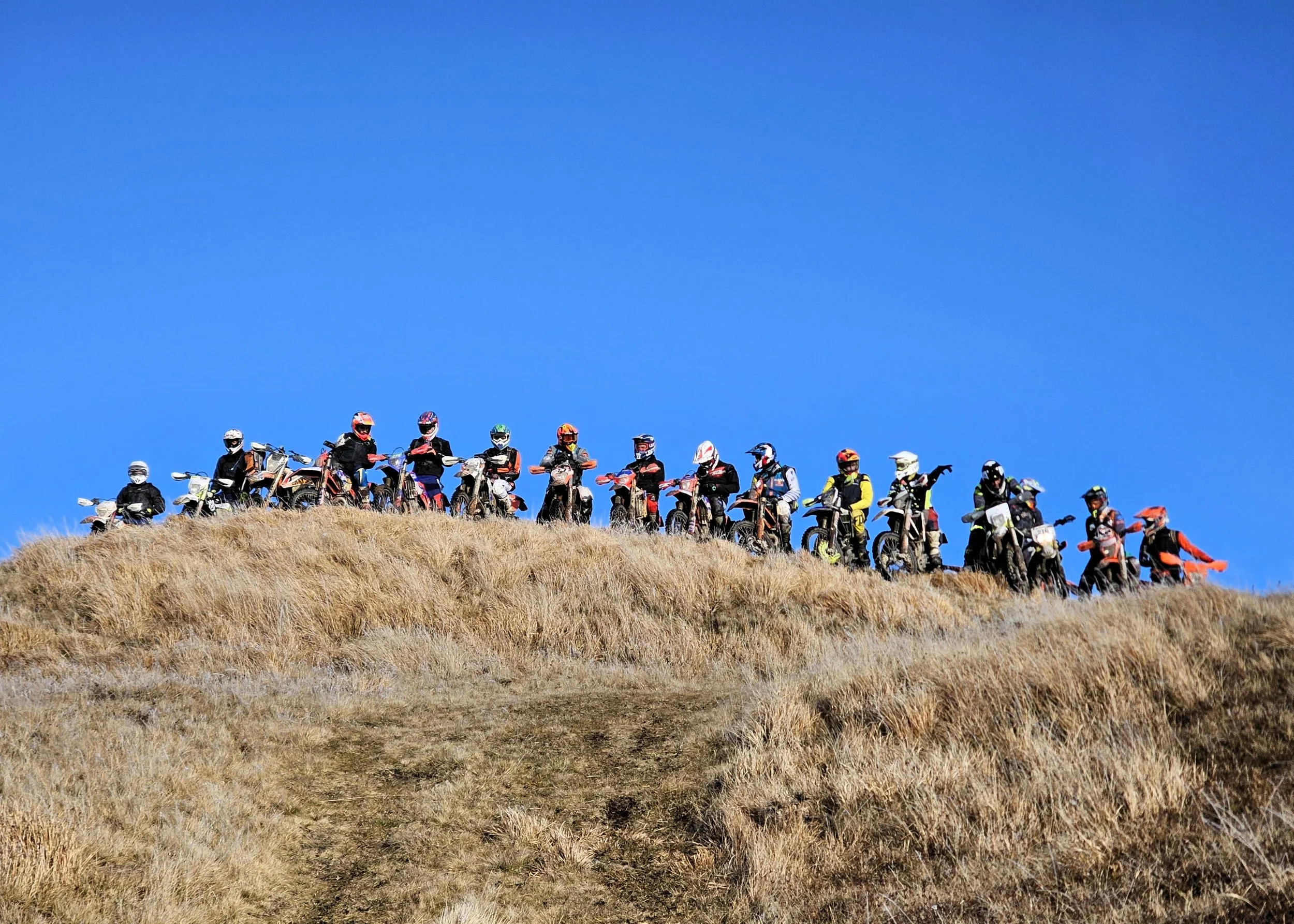 Un gruppo di motociclisti con casco e abbigliamento protettivo si trova su una collina di erba secca, sotto un cielo azzurro.