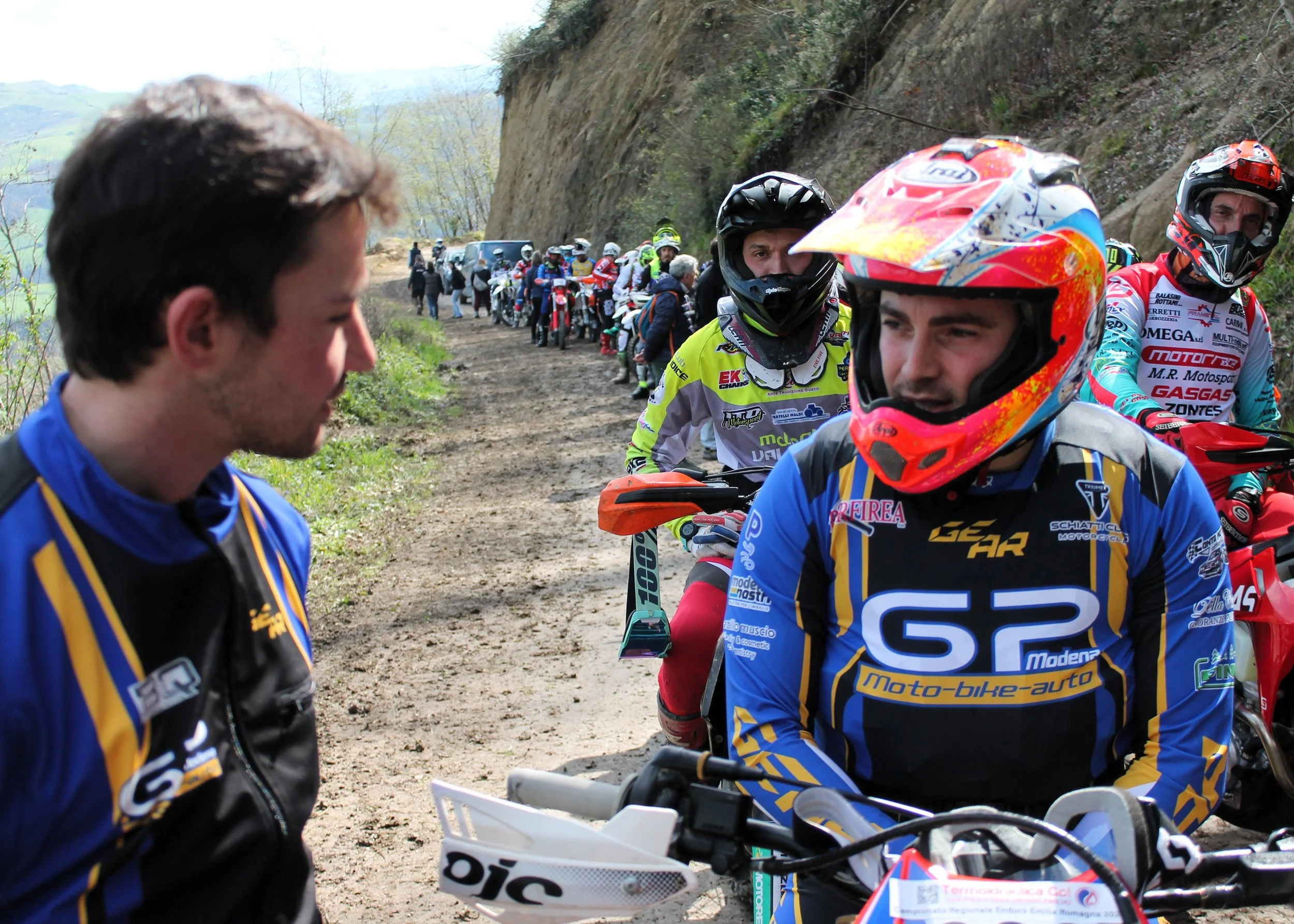 Giovani motociclisti in una gara su una strada sterrata, con uno in primo piano con casco arancione e giubbotto blu, mentre altri in background sulla strada affollata di partecipanti.