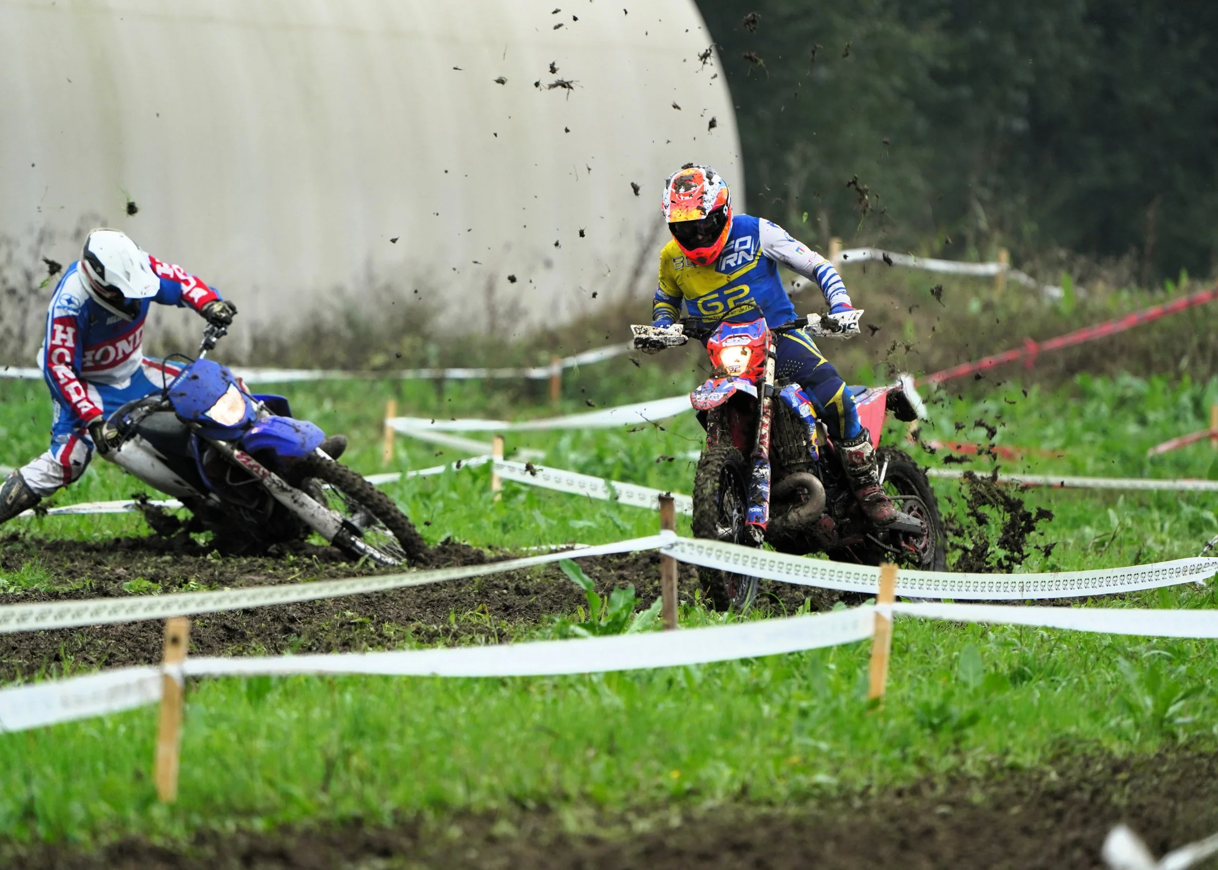 Due motociclisti gareggiano su un percorso fangoso e erboso, con uno che cade e l'altro che si mantiene in equilibrio, mentre si sollevano zolle di terra e fango.