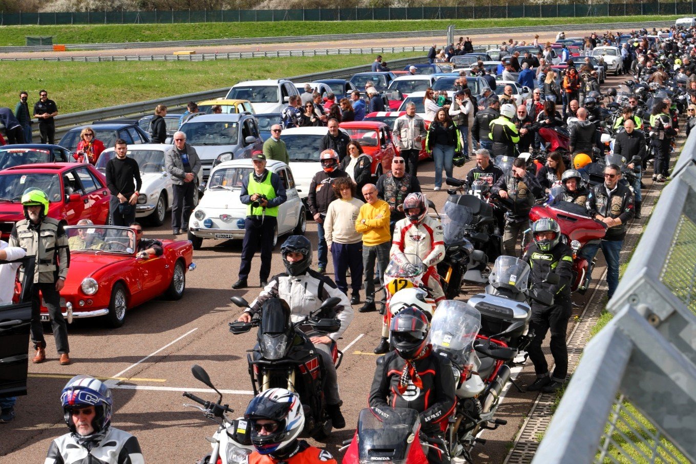 Raggruppamento di molte persone con veicoli, tra cui motociclette, scooter e auto, al bordo di una pista o area di gara, con alcuni individui in abbigliamento da motociclismo.