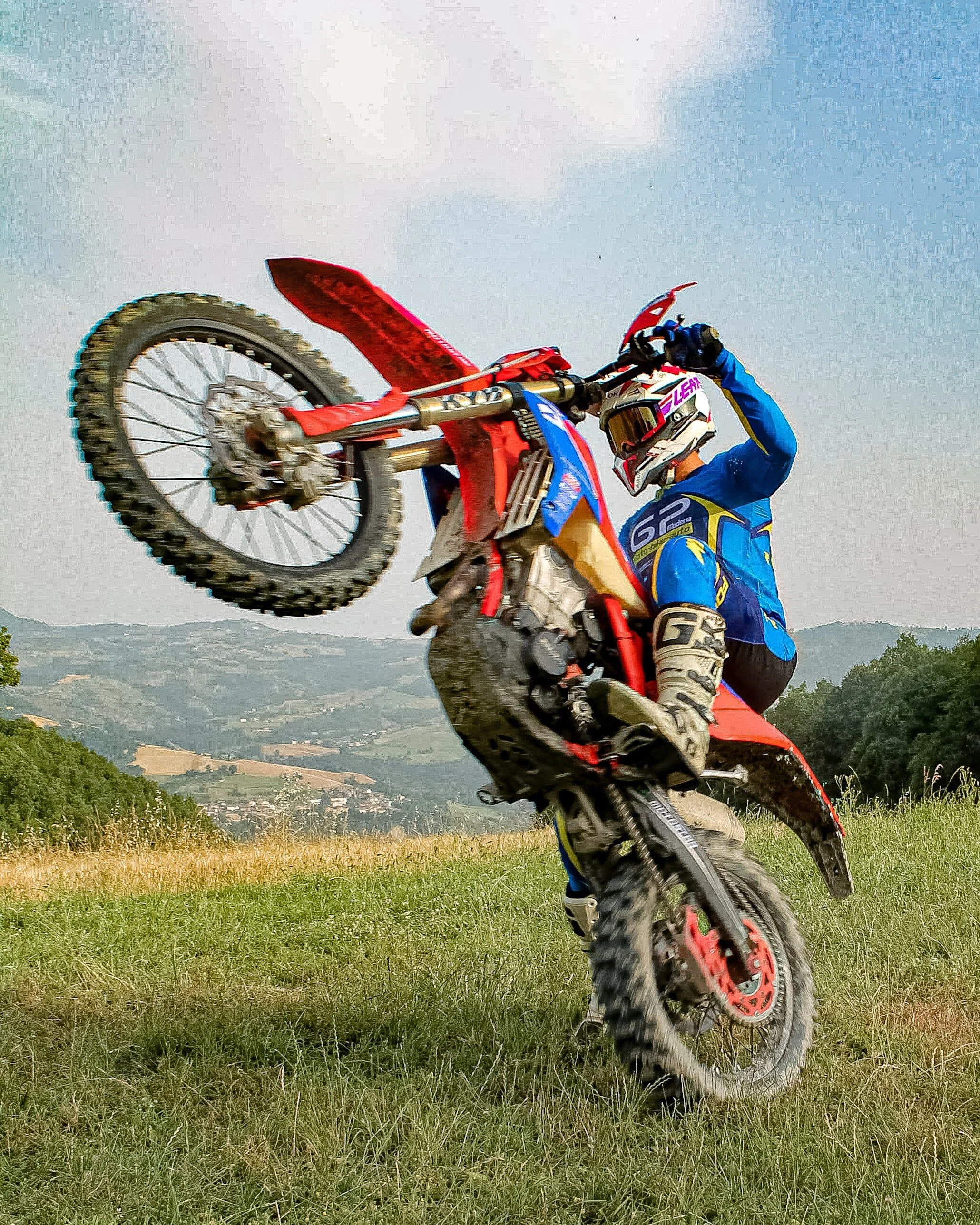 Pilota di motocross in acrobazia su una moto da cross che si solleva da terra in un campo verde con colline sullo sfondo.