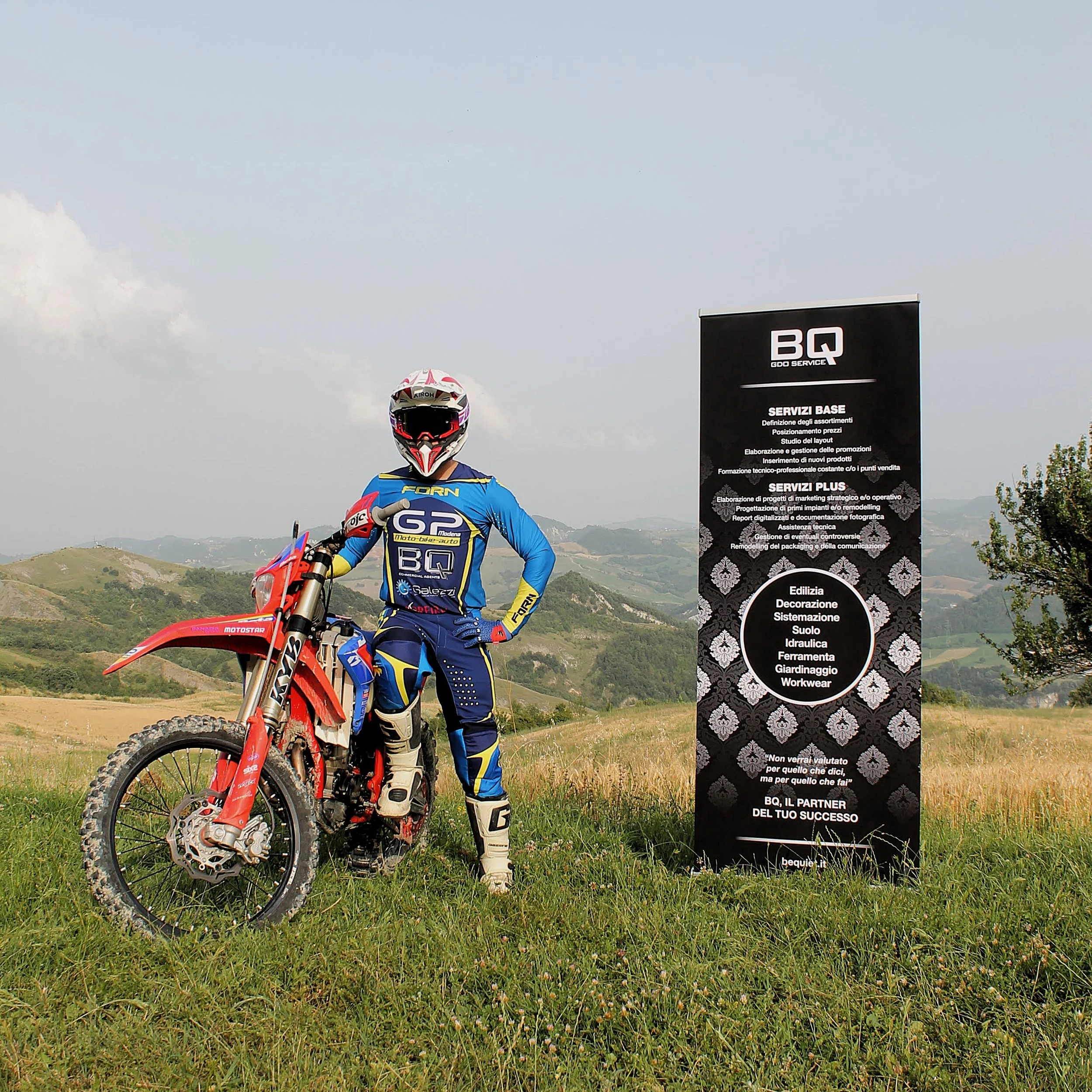 Un motociclista in abbigliamento da off-road con casco, guanti e stivali, posa accanto a una moto da cross rossa in un paesaggio collinare. C'è anche un grande cartellone nero con testo pubblicitario accanto a lui.