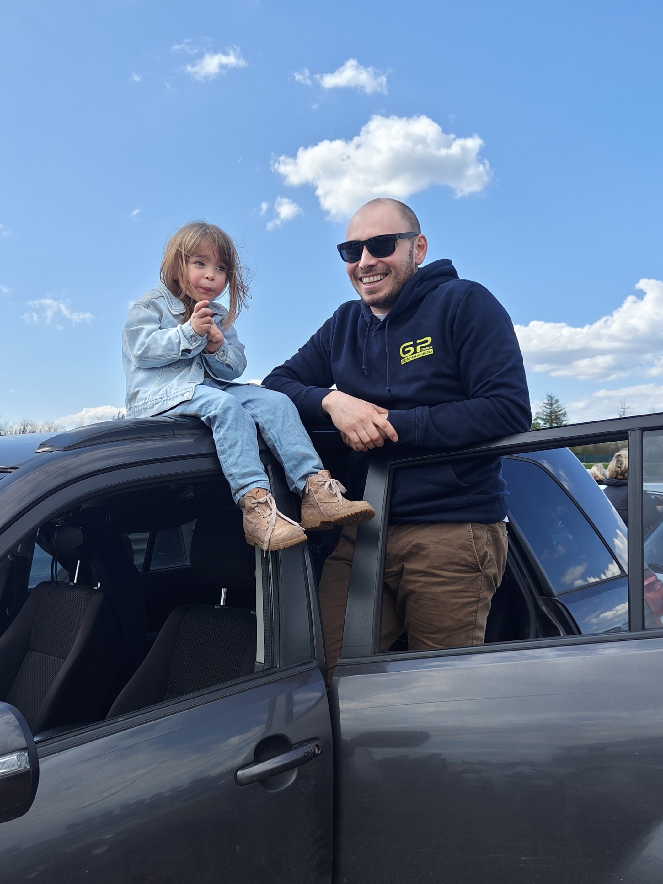 Presidente GP Modena Giuseppe Goldoni sono su una macchina, con il cielo sereno sullo sfondo. La bambina è seduta sul tetto dell'auto e l'uomo la tiene per mano, entrambi sorridenti.