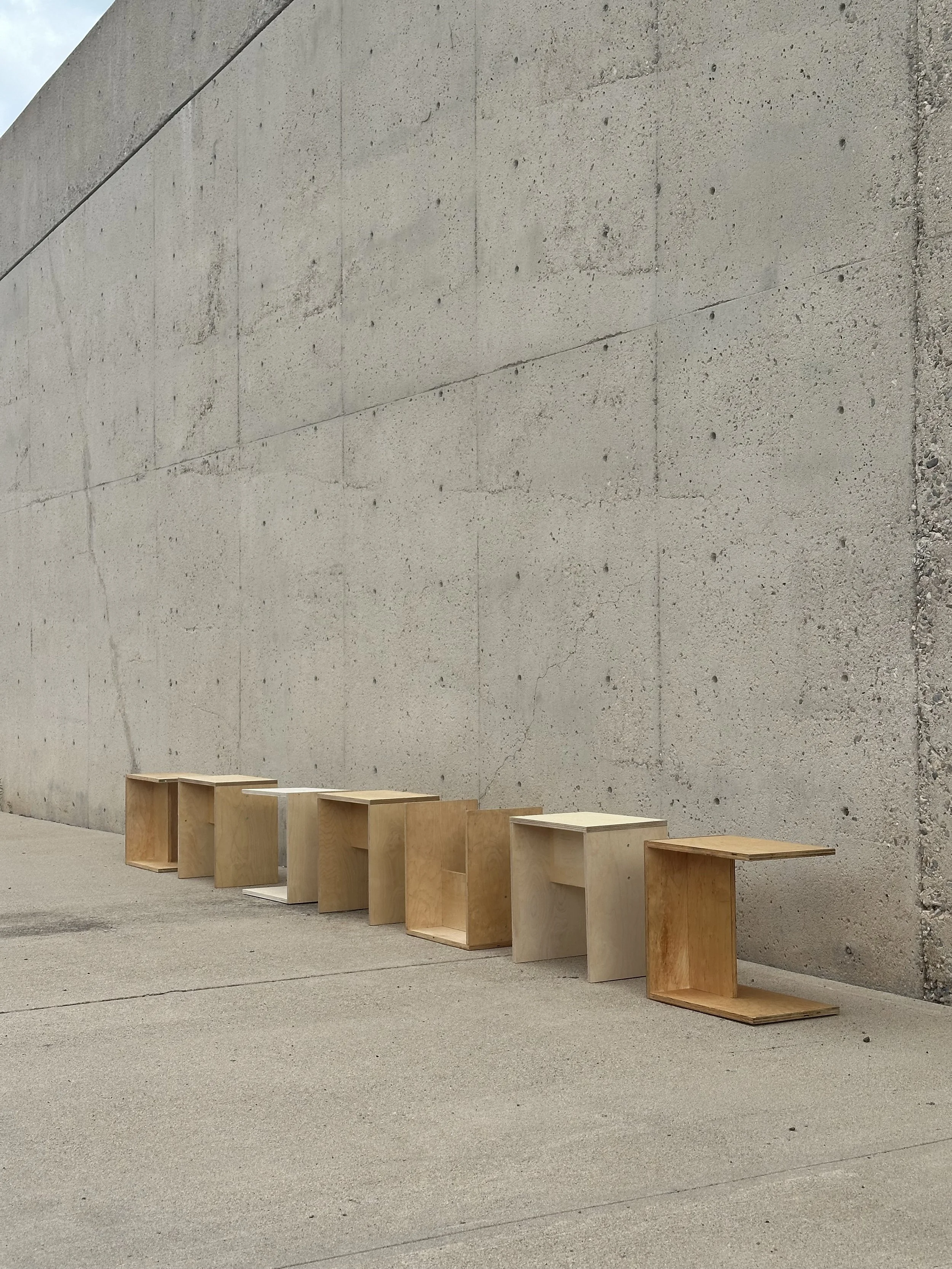 A row of wooden benches with open sides and minimalistic design, placed against a tall concrete wall outdoors.
