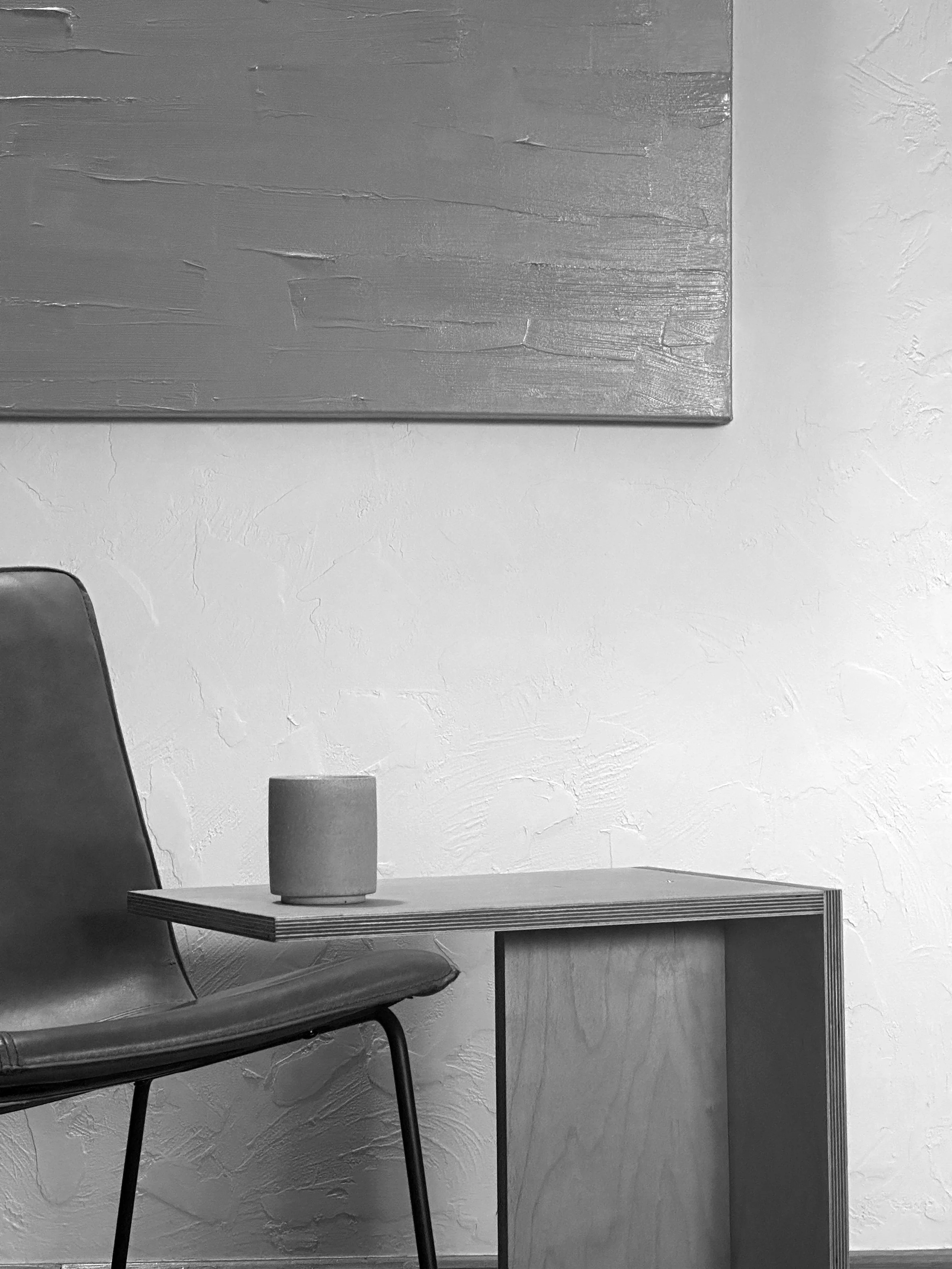 A black and white photo of a modern interior space showing part of a chair, a small table with a cup or mug, a textured wall, and a wooden art piece on the wall.