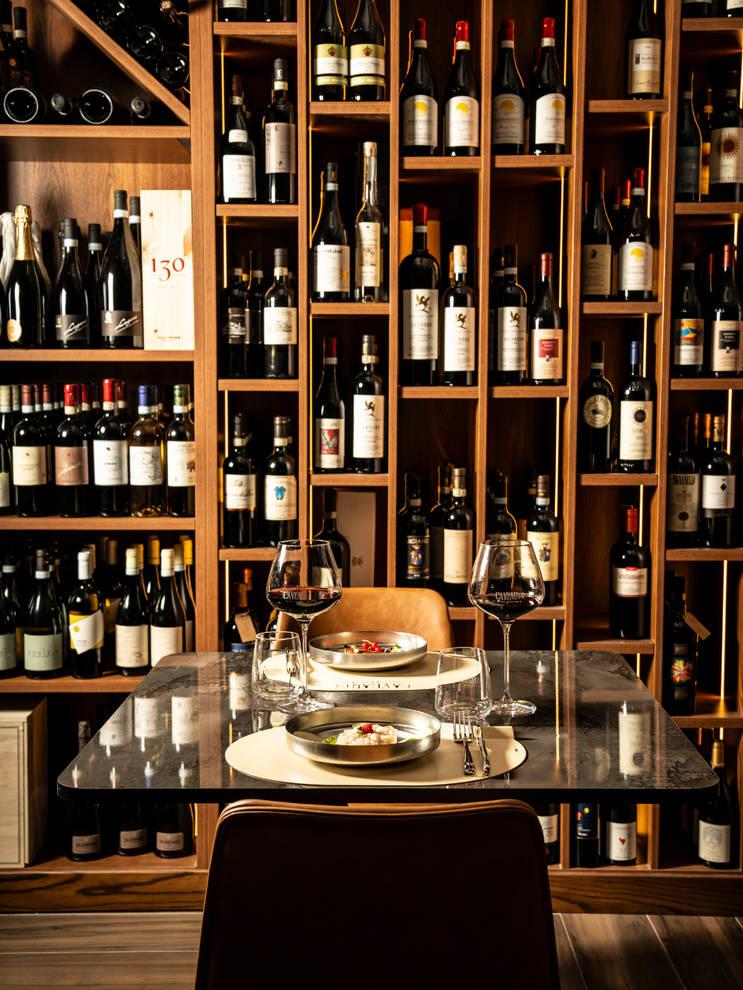 Tavola apparecchiata con due bicchieri di vino rosso, piatti con cibo, posate e bicchieri d'acqua, davanti a una parete di scaffali di vino in una enoteca o ristorante elegante.