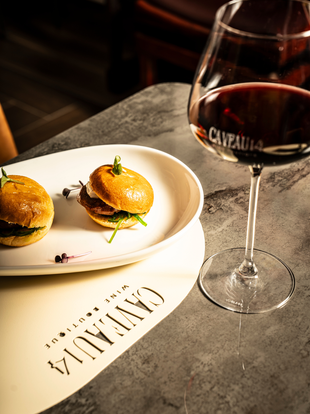 Piccola porzione di due mini burger con carne, insalata e salsa, serviti su un piatto di ceramica con scritta "CAVALIERI WINE & FOOD" accanto a un bicchiere di vino rosso.