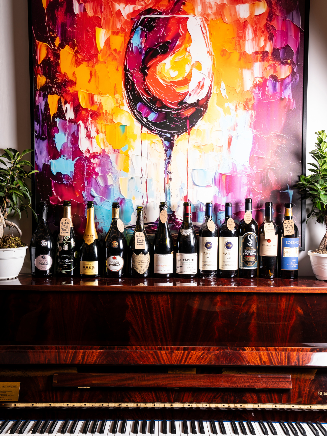 Un quadro astratto colorato con un bicchiere di vino rosso stilizzato sopra una series di bottiglie di vino su un mobile in legno, con due piante verdi ai lati.