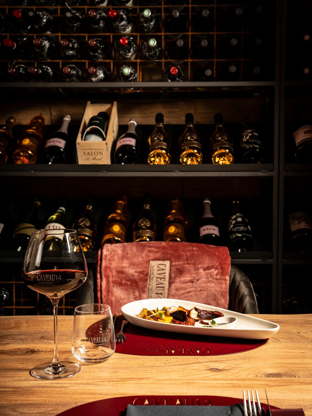 Una tavola con un bicchiere di vino rosso, un piatto di cibo, e un bicchiere vuoto, in un ambiente di ristorante con scaffali di bottiglie di vino sullo sfondo.