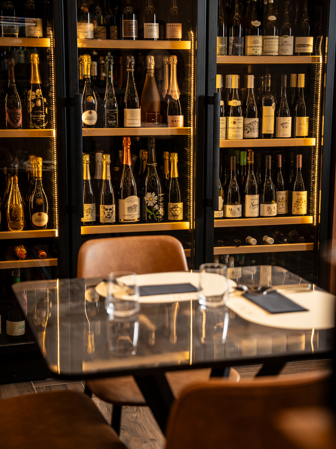 Vetrina frigorifera con bottiglie di champagne e vino, tavolo apparecchiato con bicchieri e piatti nel ristorante.