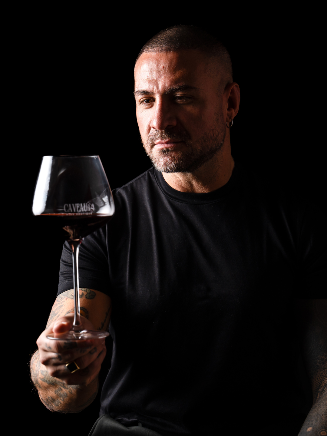 Uomo con tatuaggi che tiene un bicchiere di vino rosso, sfondo nero.