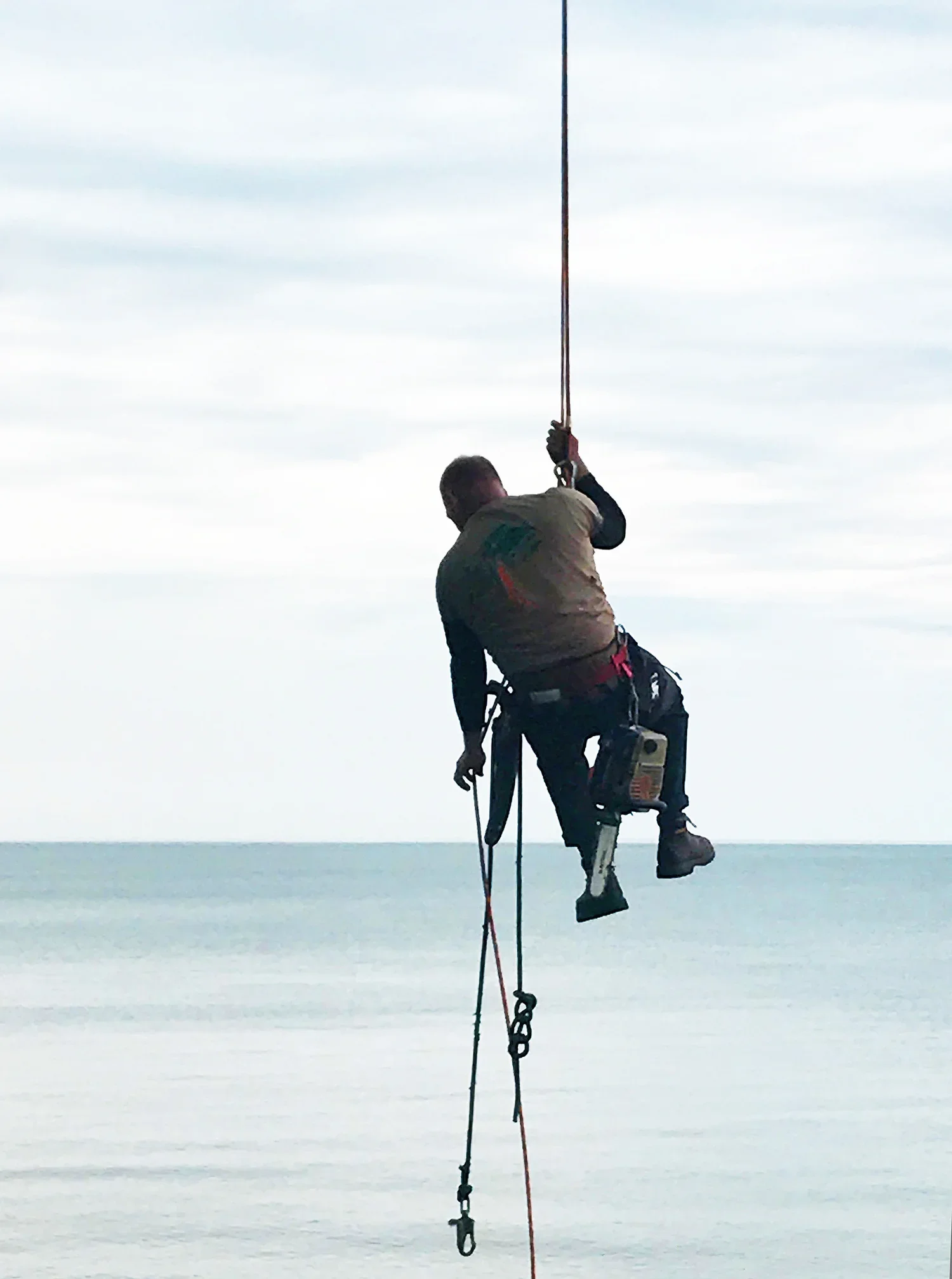 A person wearing climbing gear hanging from a rope over water, with a cloudy sky in the background.