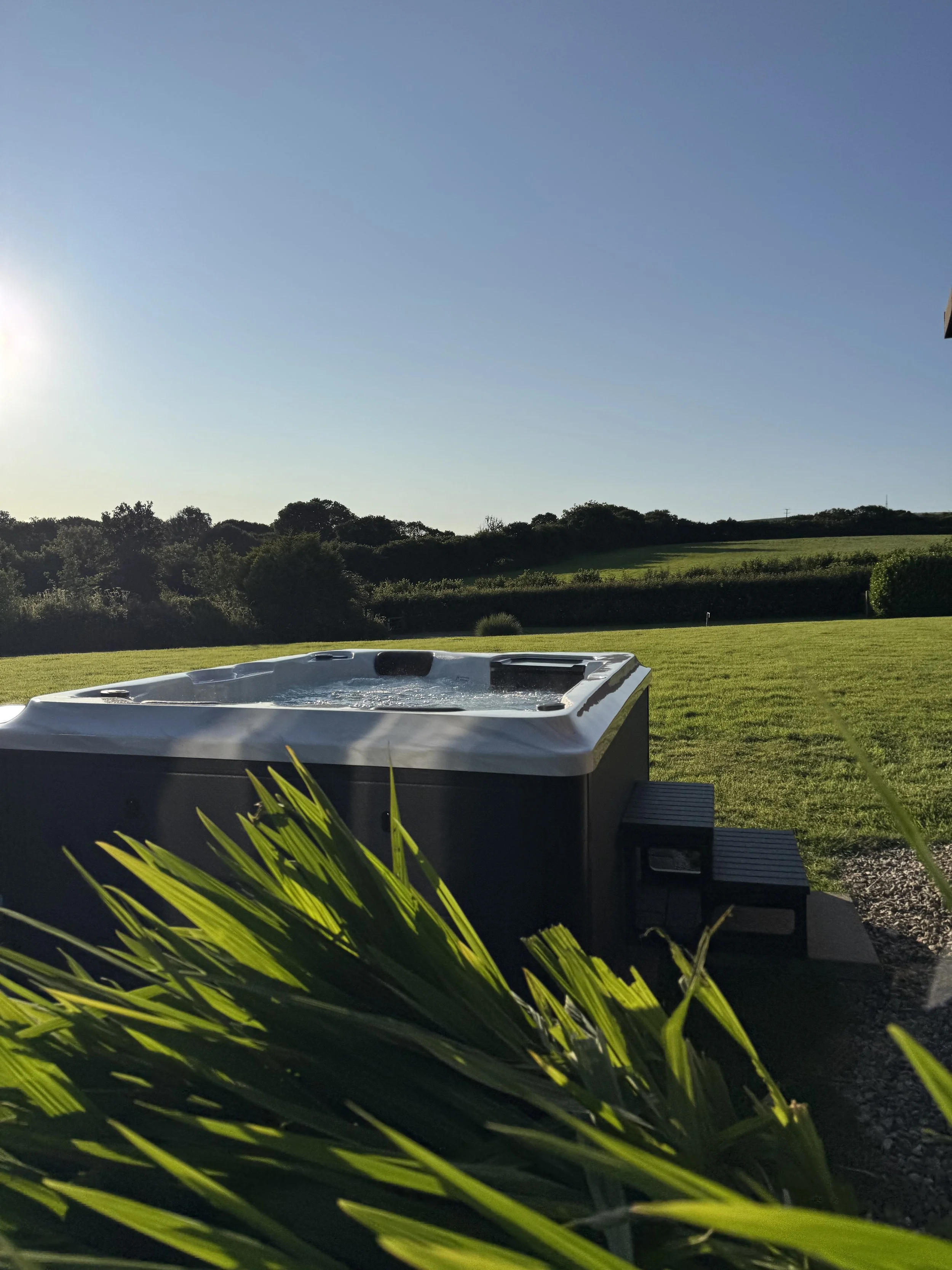 Hot tub outside a pod, looking out over grass up onto green hills and trees in the background during sunset.