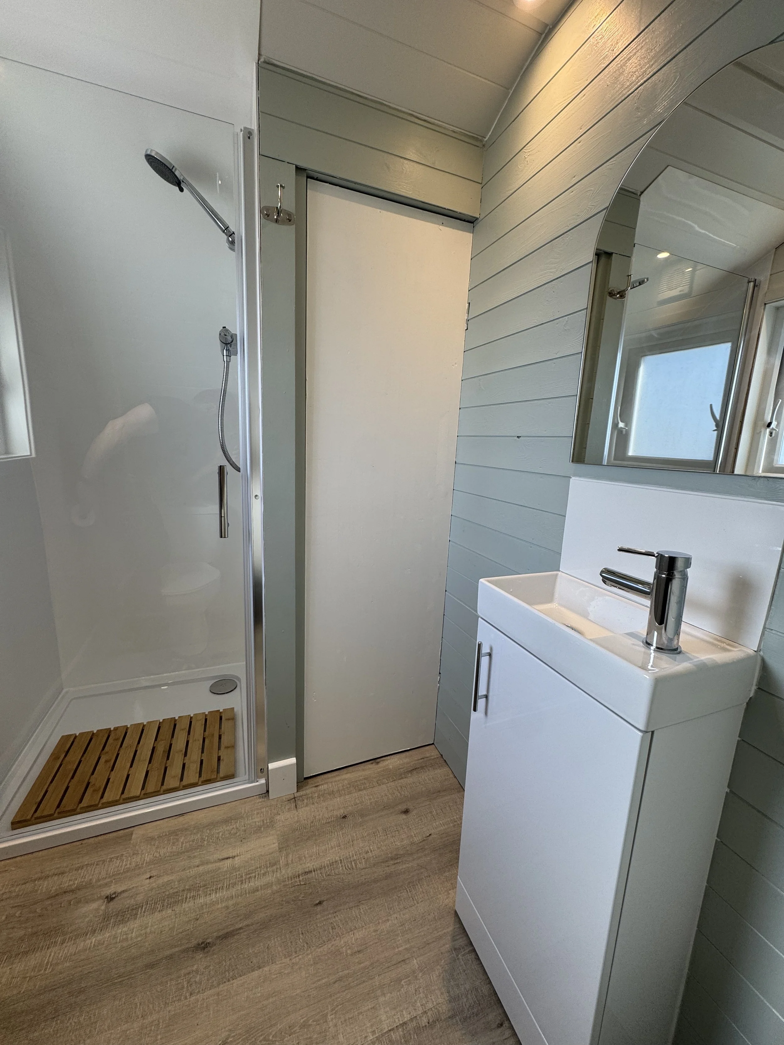 A small bathroom featuring a shower with a glass door, a wooden shower mat, a white sink with a modern chrome faucet, a light-colored wooden floor, and a wall with horizontal light blue paneling.
