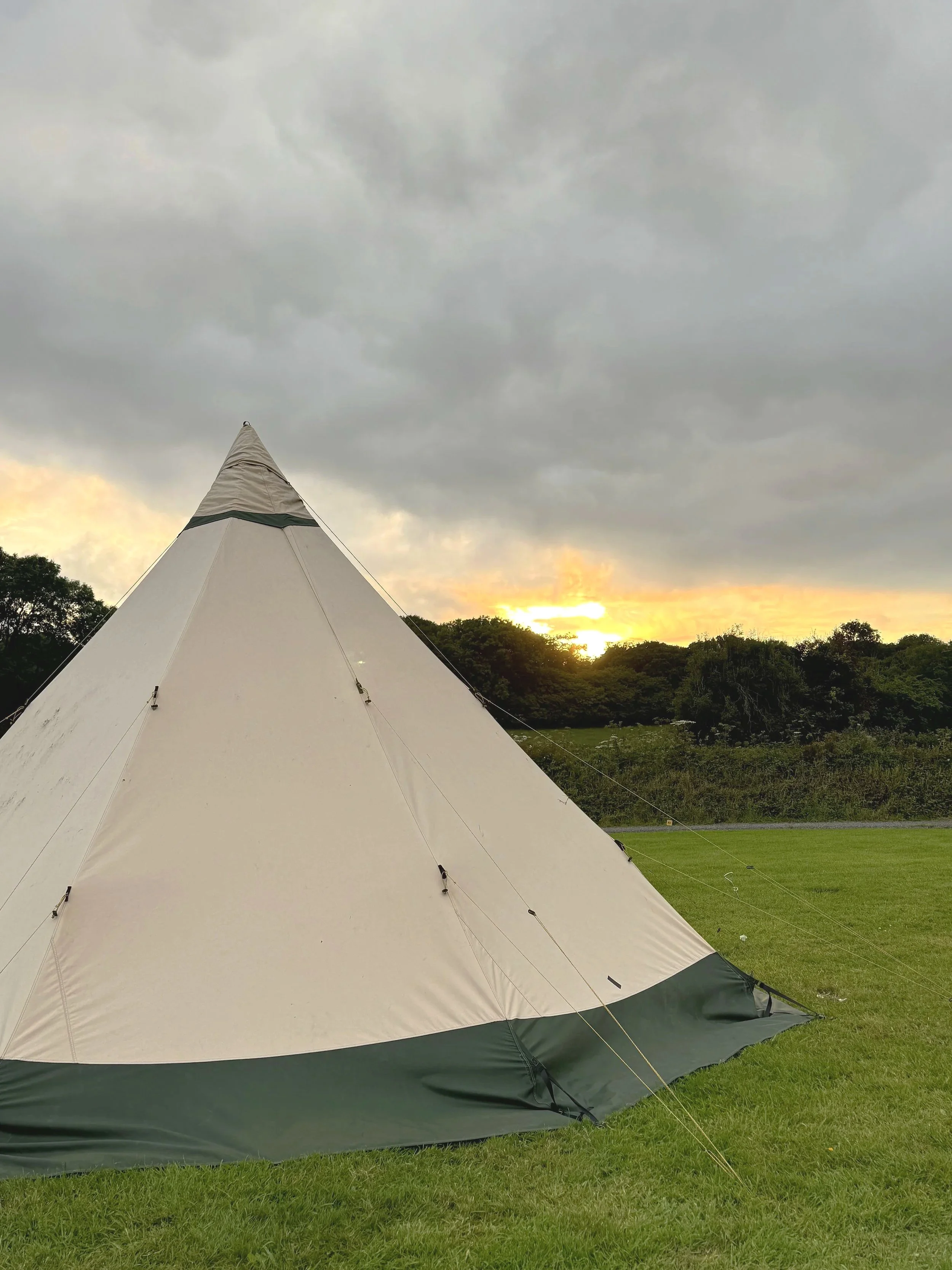 A beige camping bell tent with a green base, set up on a grassy field, with a backdrop of trees and a cloudy sky during sunset.