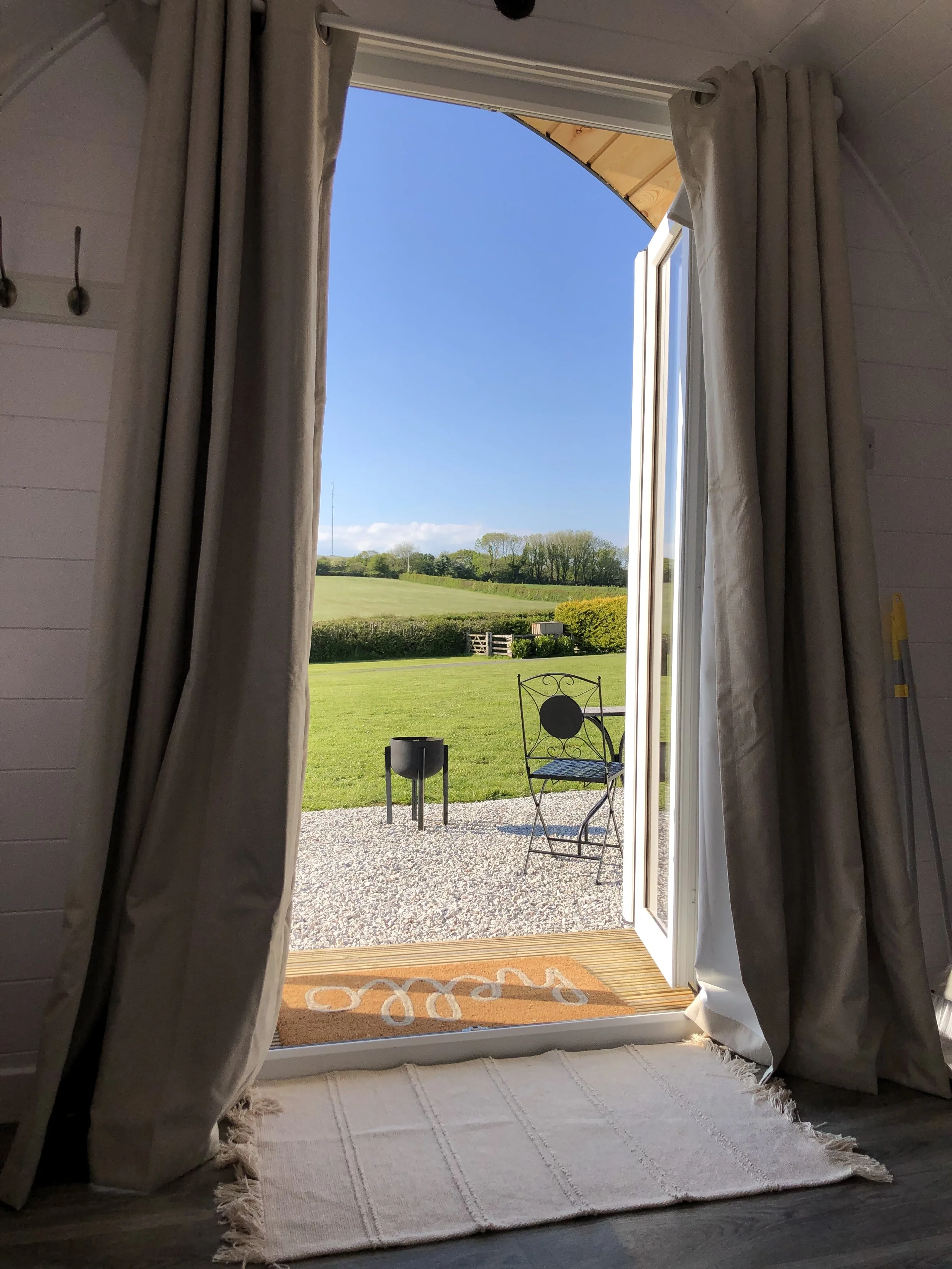 Open door leading to a patio with a metal chair and a small black fire pit, overlooking a grassy yard and green fields under a clear blue sky.