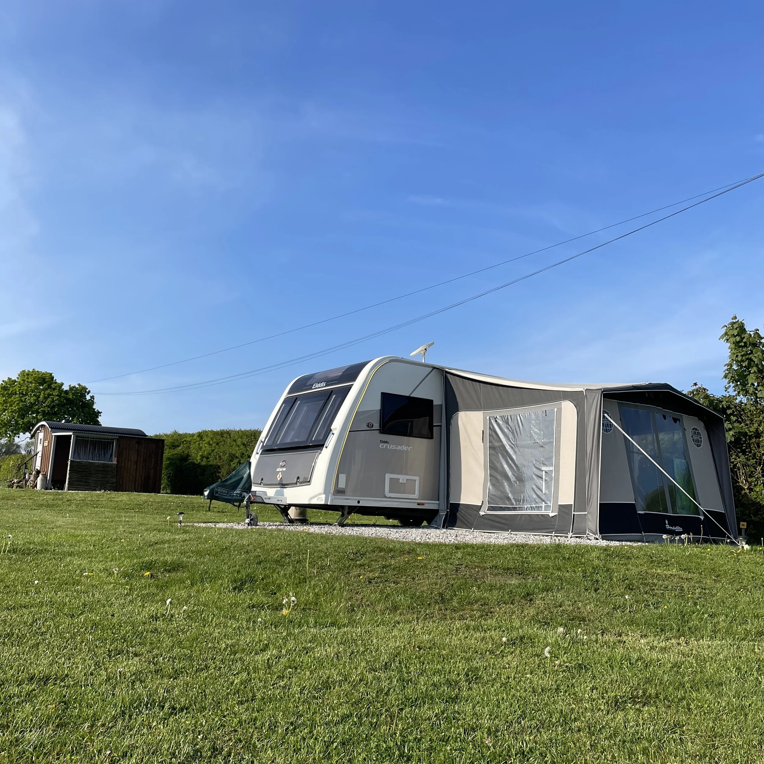 A caravan with an awning set up on a grassy field under a clear blue sky, with a small wooden shepherds hut and trees in the background.