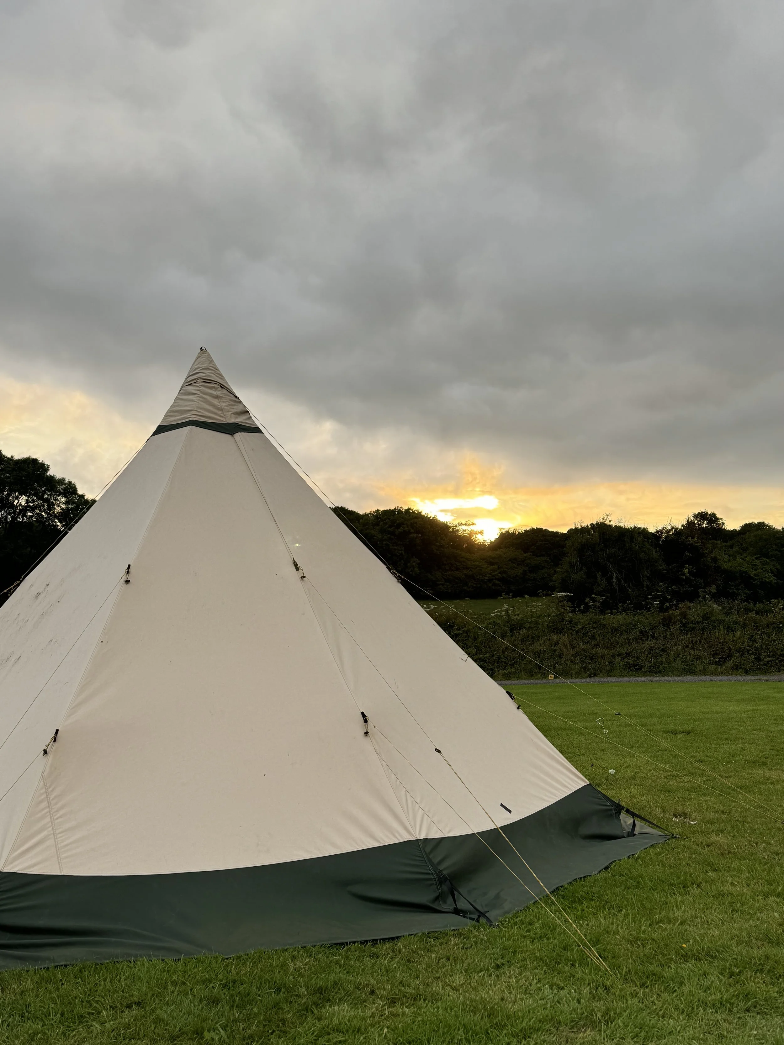 A beige camping bell tent with a green base, set up on a grassy field, with a backdrop of trees and a cloudy sky during sunset.