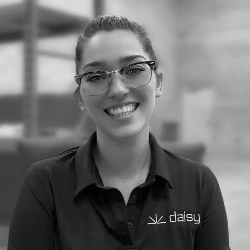 A young woman with glasses and a smiling expression posing for a photo, wearing a collared shirt with a logo and the word 'daisy' embroidered on it.