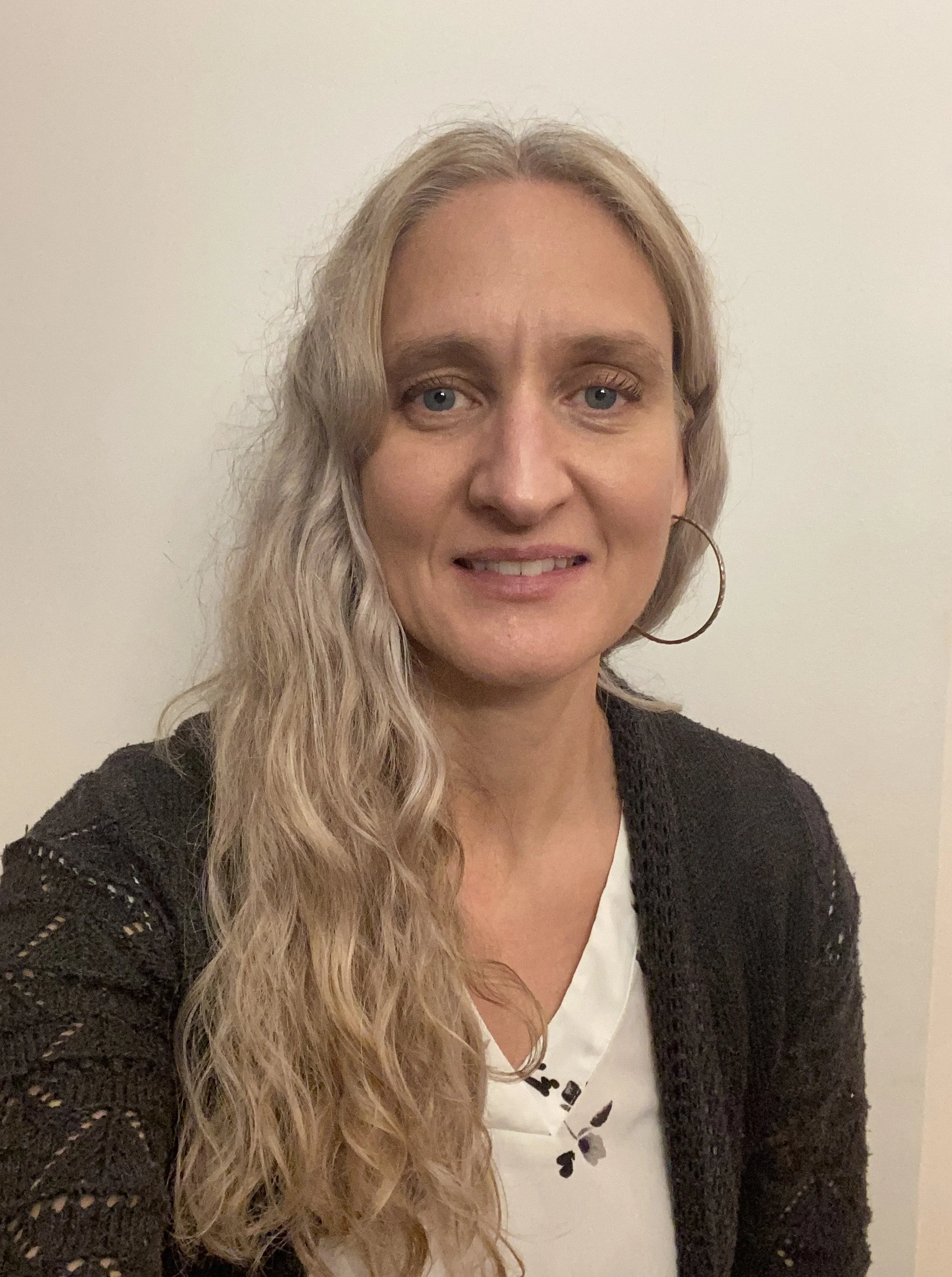 Une femme avec de longs cheveux blonds bouclés, portant un pull noir en crochet, une chemise blanche avec des motifs violets et des boucles d'oreilles en forme de cercle, posant devant un fond blanc.