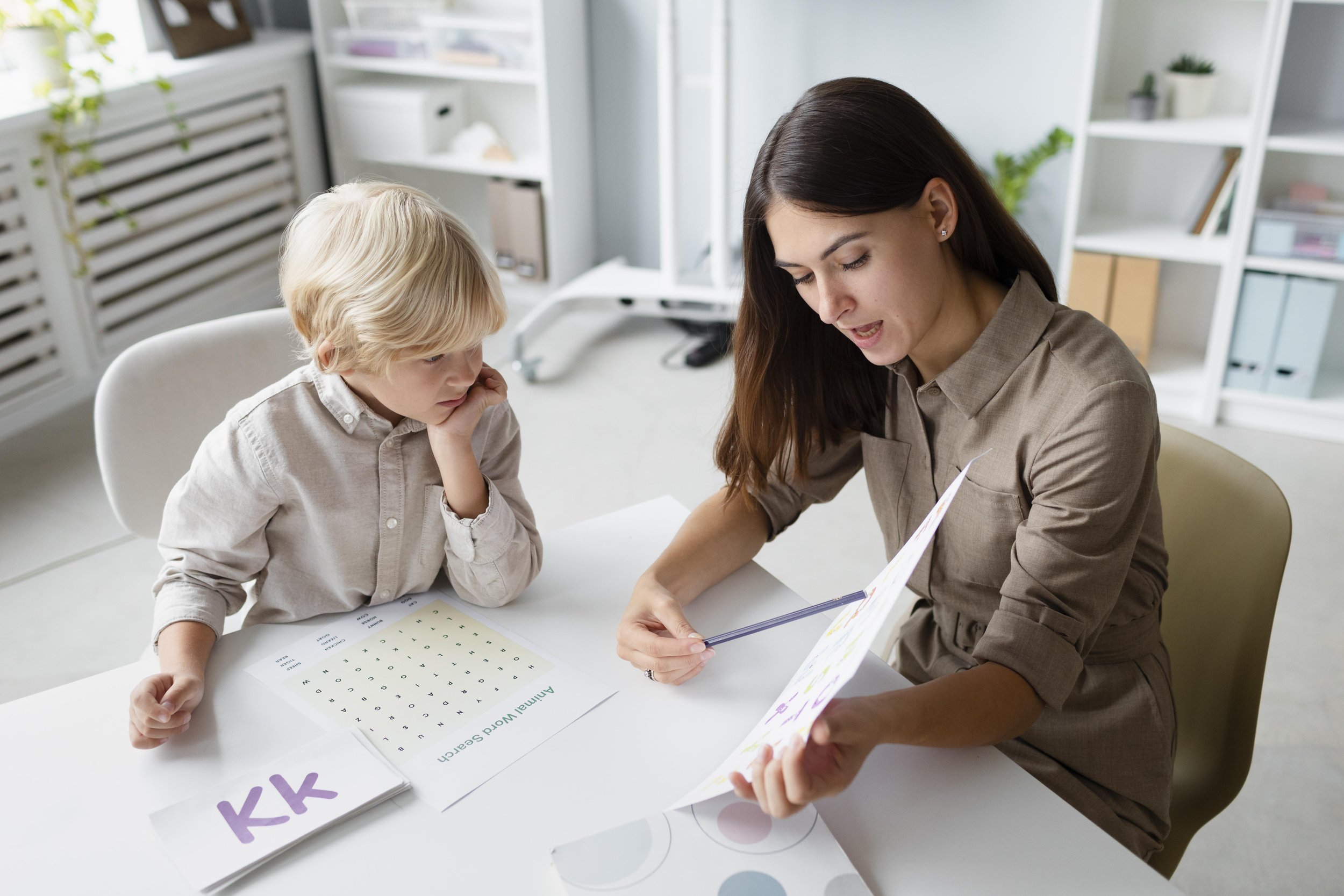 Une femme et un jeune garçon assis à une table d'apprentissage, la femme montre un document coloré à l'enfant, fond d'une pièce lumineuse avec des étagères blanches et des plantes.