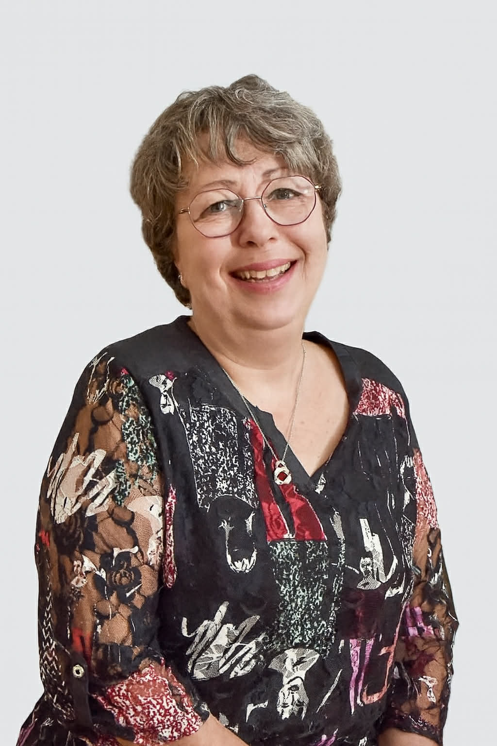 Une femme souriante portant des lunettes, avec des cheveux courts et bouclés, habillée d'une blouse à motifs colorés, de fond blanc.