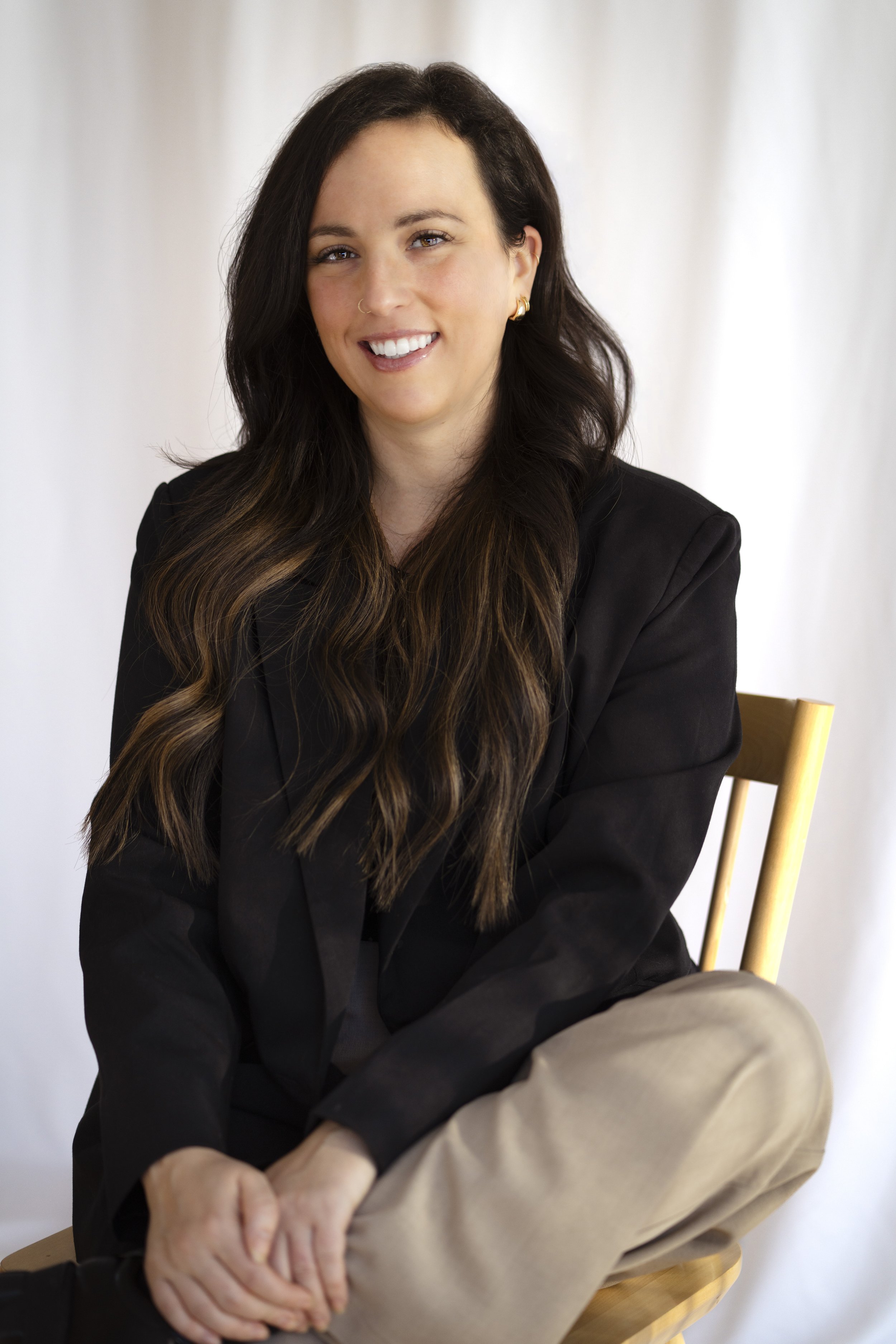 Une femme assise sur une chaise en bois, souriante, avec de longs cheveux bruns ondulés, portant une veste noire et un jean beige, contre un fond blanc.