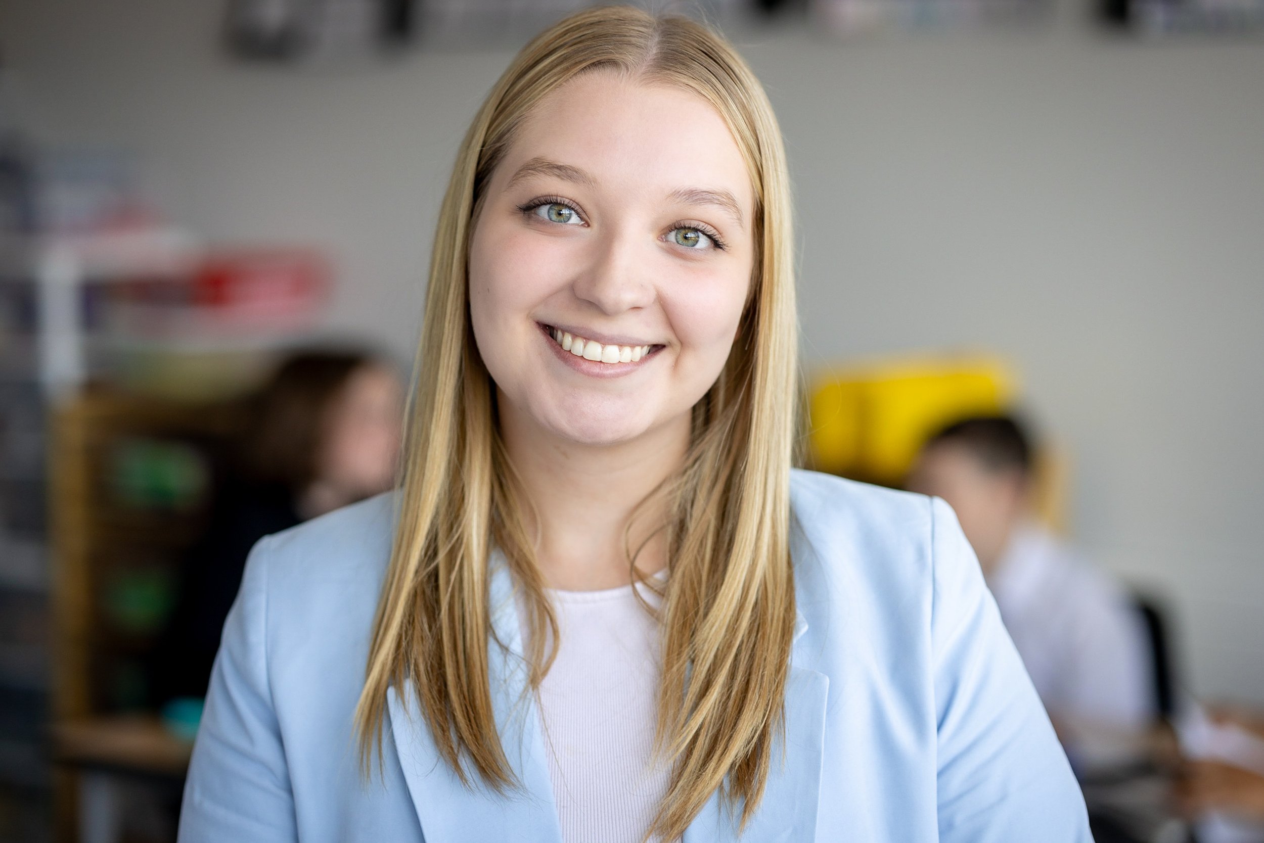 Une jeune femme blonde souriante portant une veste bleue dans un environnement de bureau ou d'école, avec d'autres personnes floues en arrière-plan.
