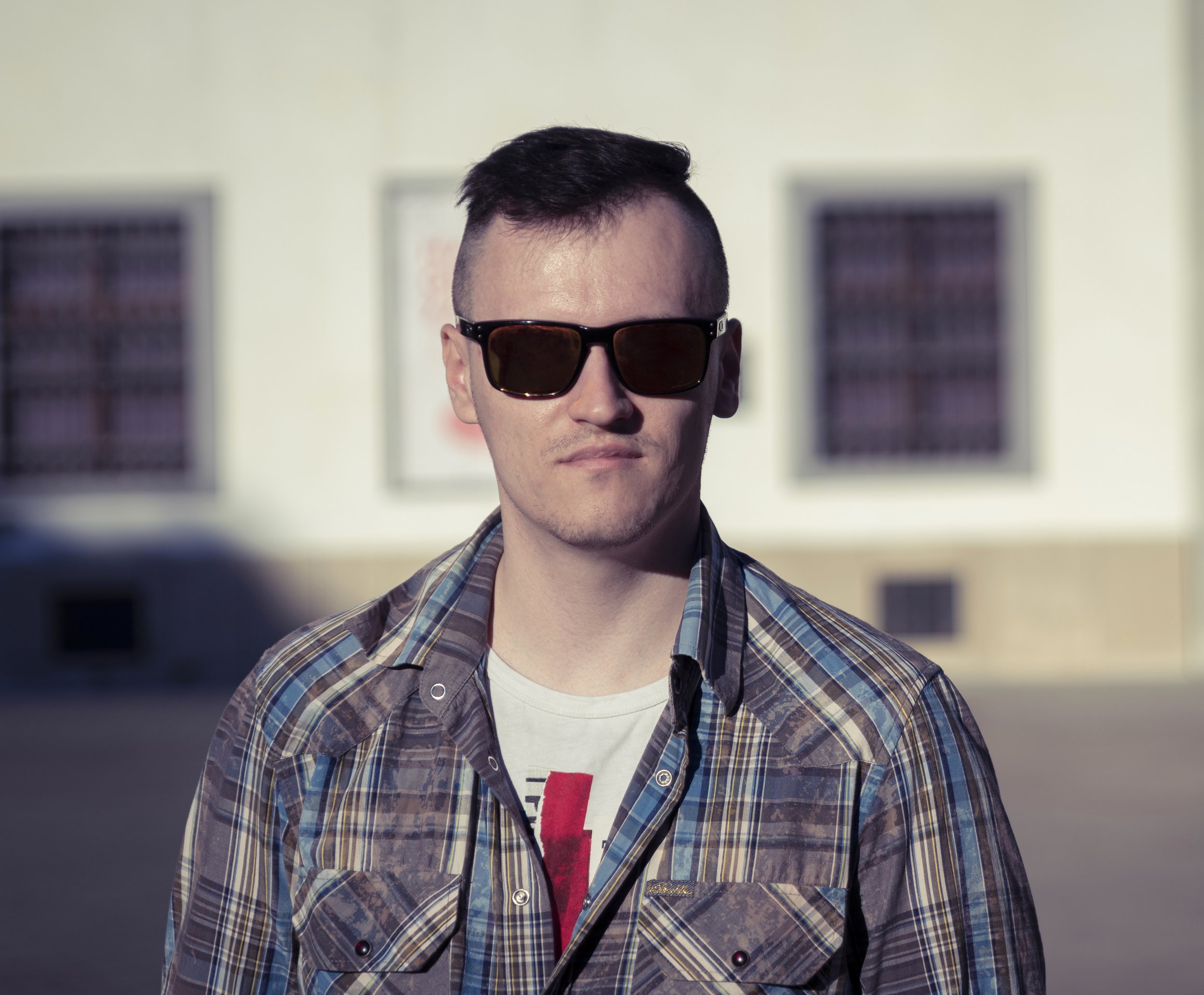 A young man with short dark hair wearing sunglasses, a plaid shirt, and a graphic T-shirt, standing outdoors in front of a blurred building background.