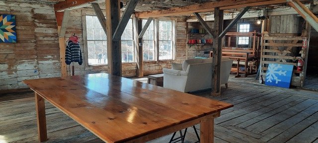 Rustic interior of a wooden cabin with a large wooden table, a white armchair, windows, and shelves with books.