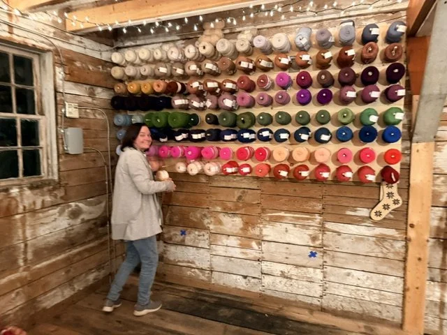 A woman in a gray coat and jeans standing next to a wall of colorful yarn balls in a rustic wooden room.