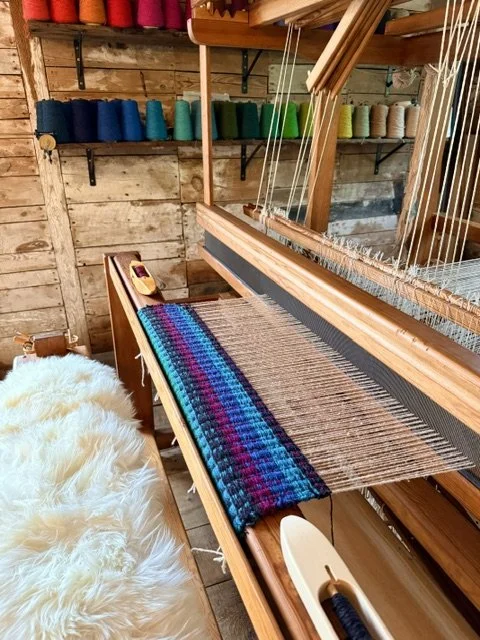 A weaving loom with a partially woven colorful blanket in progress, set in a rustic wooden workshop with shelves of yarn in various colors.